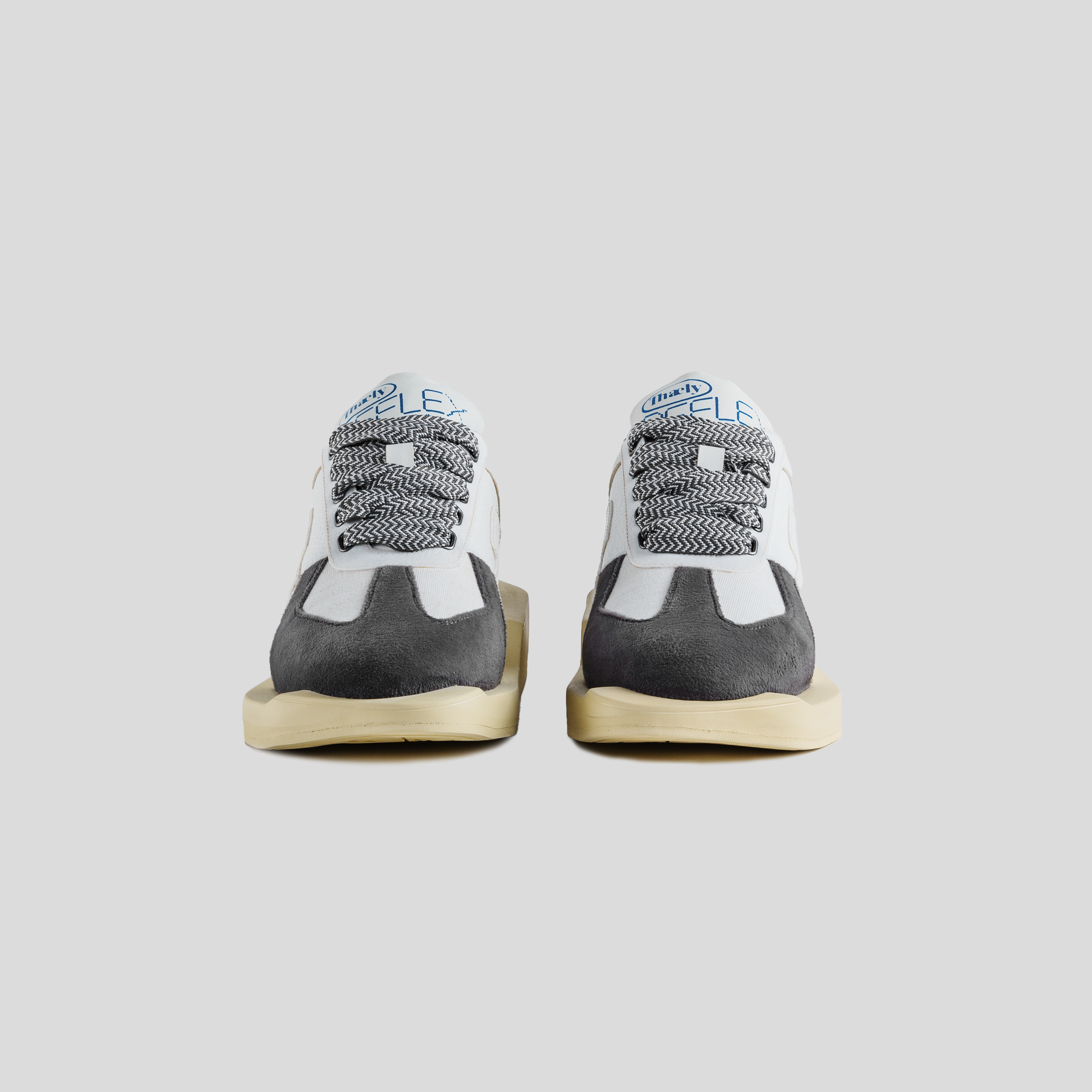 Front view of white and grey suede sneakers with black patterned laces on a neutral background.