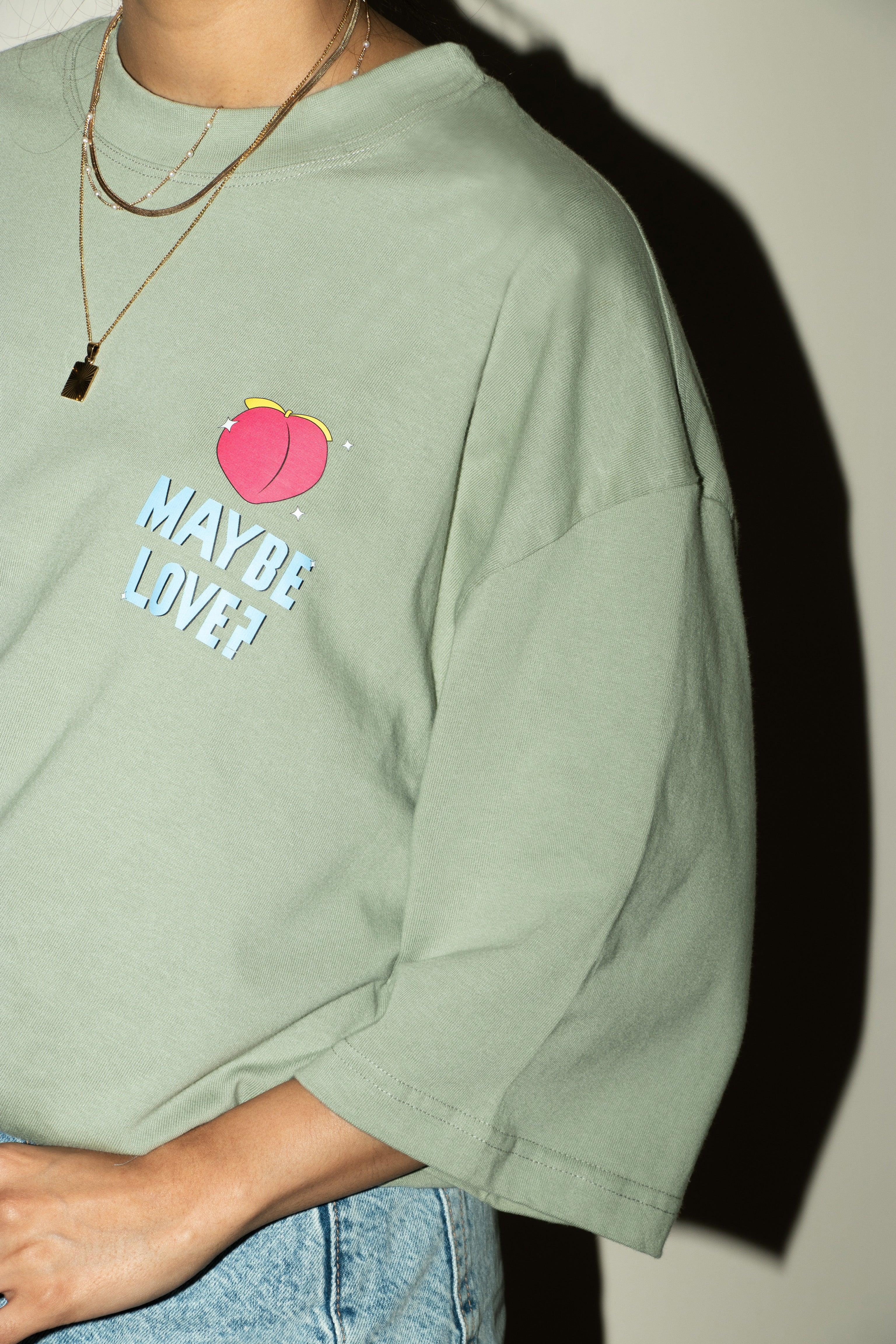 Green streetwear t-shirt with peach graphic and 'MAYBE LOVE?' text, layered gold necklaces
