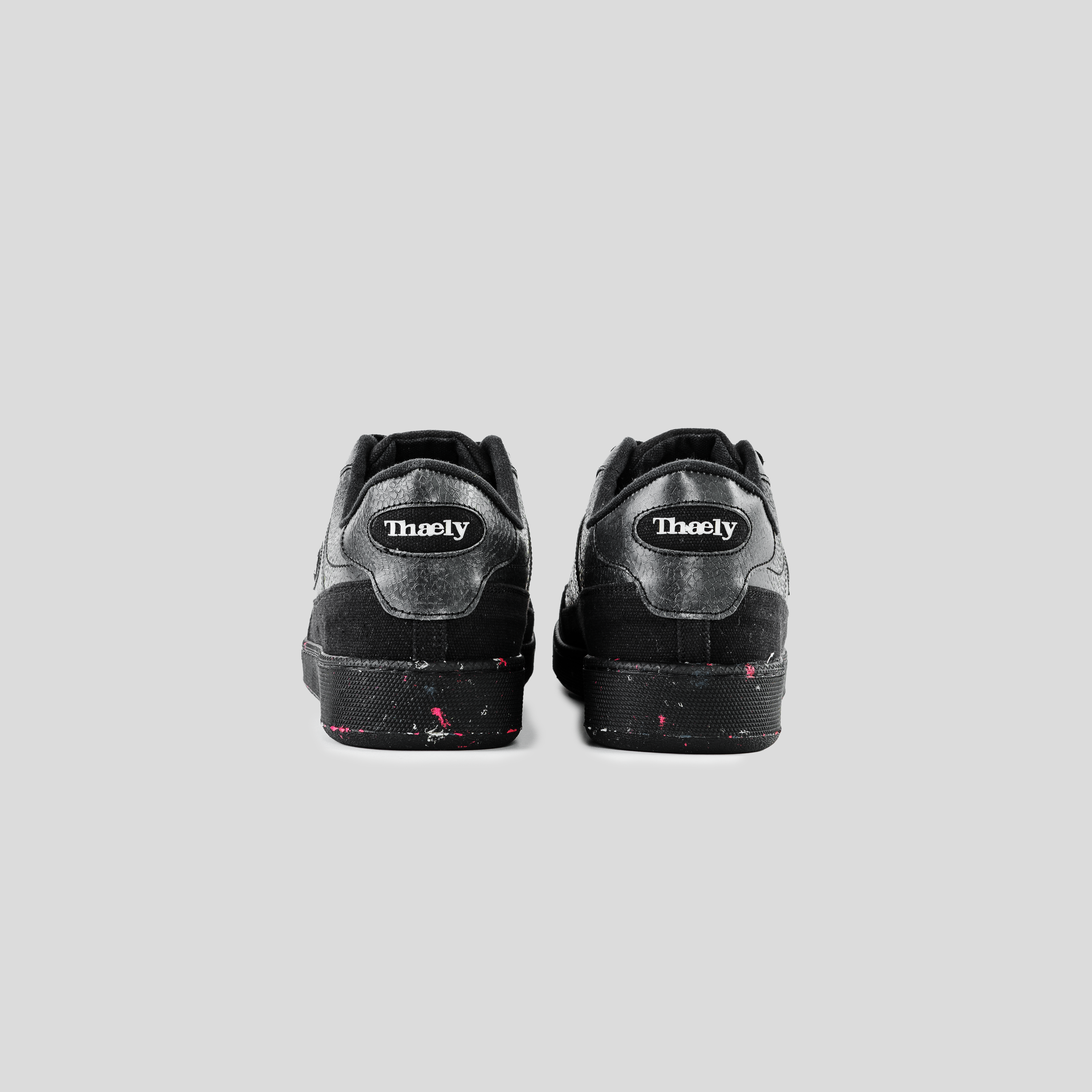 Black Thaely sneakers with white logo, pink paint splatters, rear view on gray background