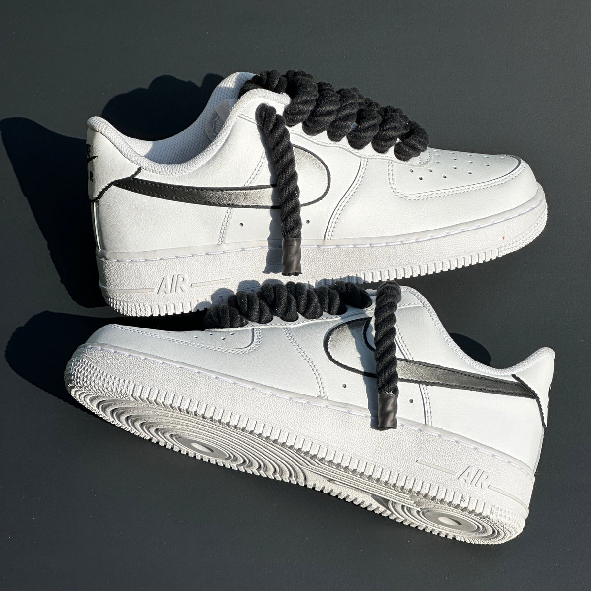White streetwear sneakers with thick black rope laces and black swoosh detail on grey background