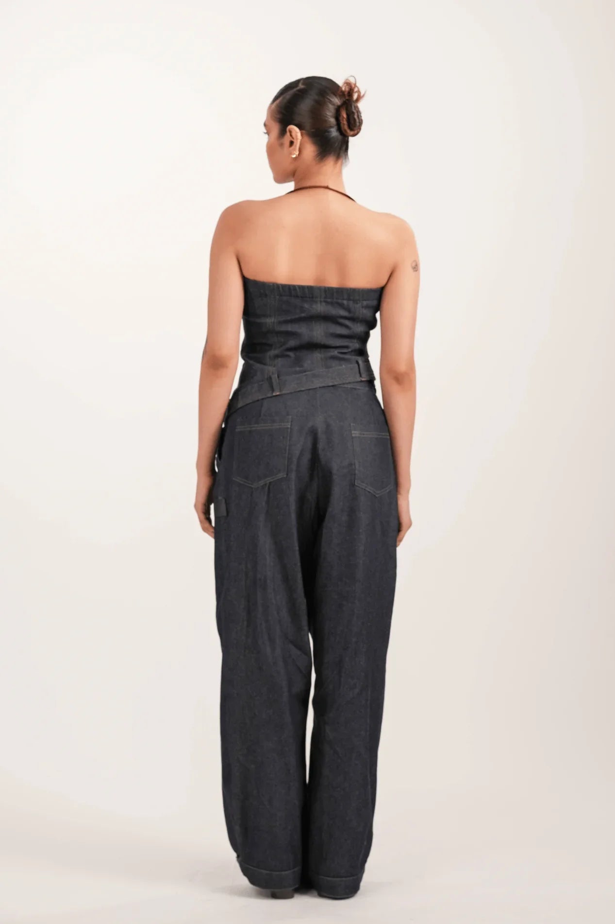 Woman wearing dark denim strapless jumpsuit with wide legs, back view against plain background