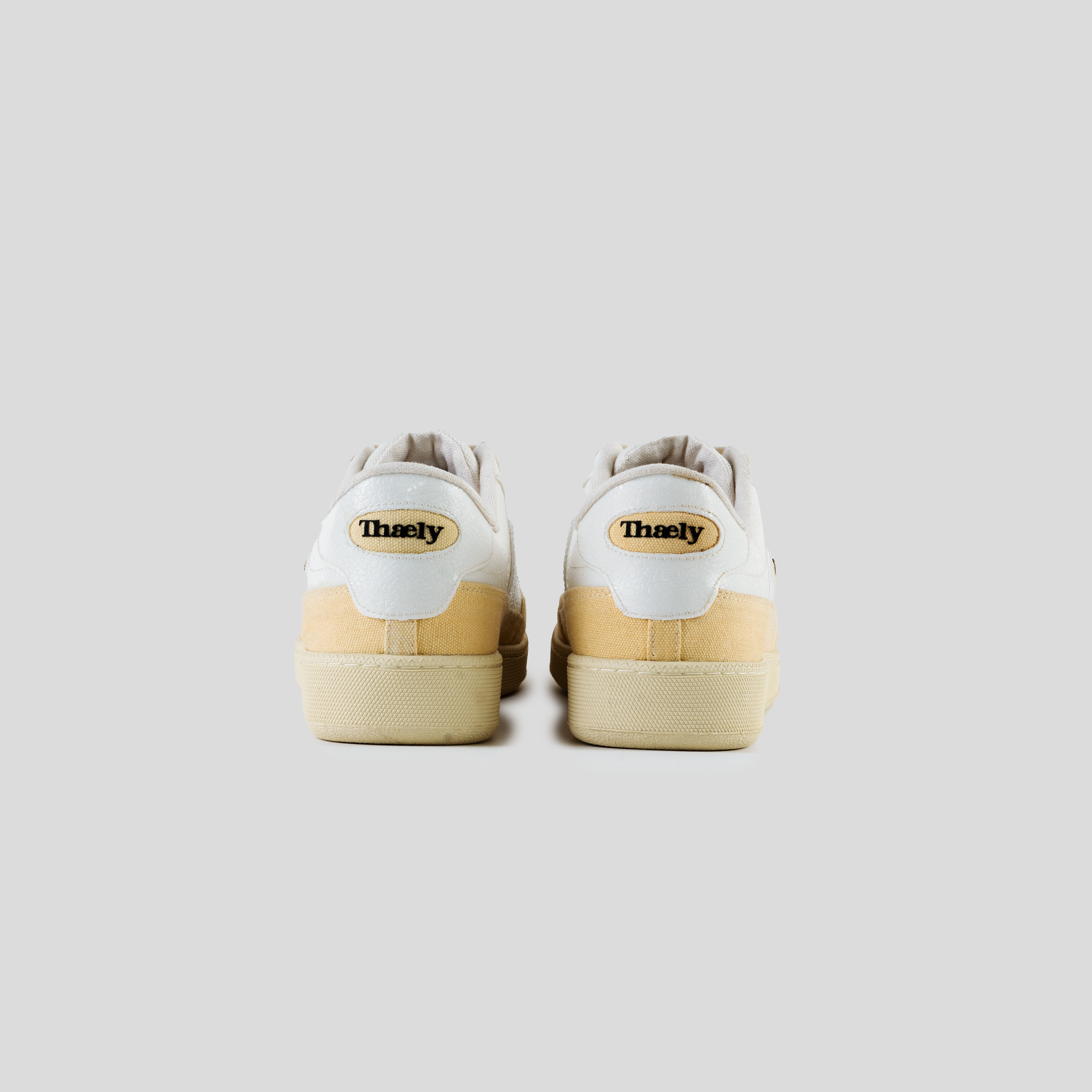 Rear view of white and beige Thaely sneakers with logo on heel, minimalist background
