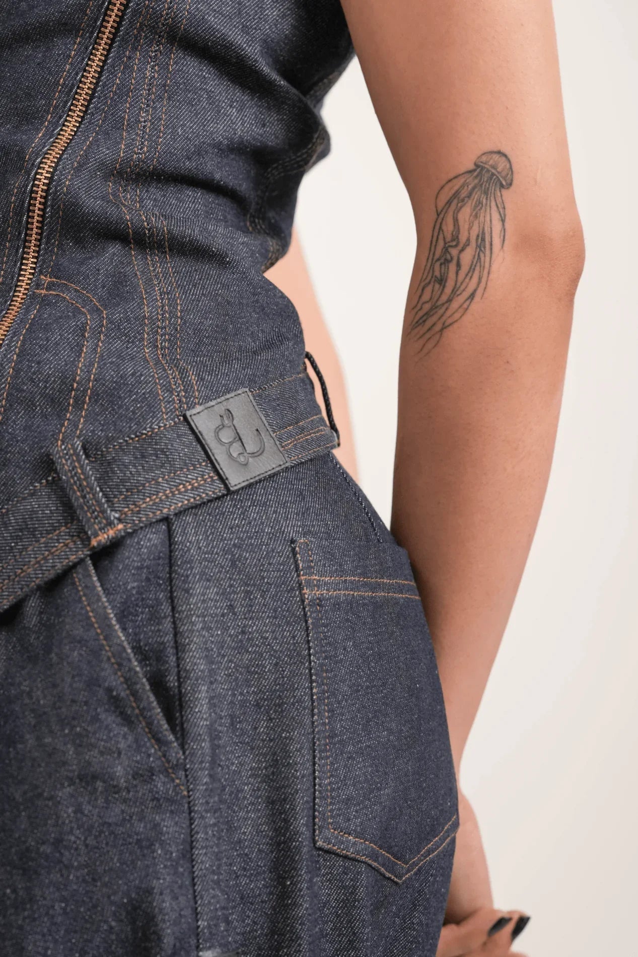 Close-up of dark denim jeans with orange stitching, side pocket, zipper and jellyfish tattoo on arm