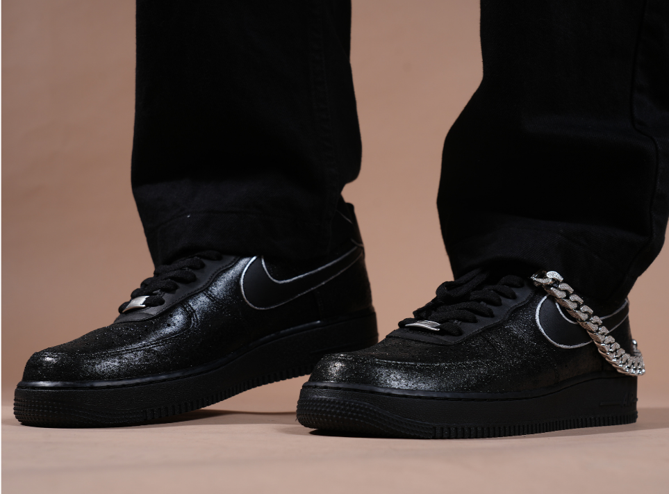 Black streetwear sneakers with metallic accents and silver chain detail, worn with black pants
