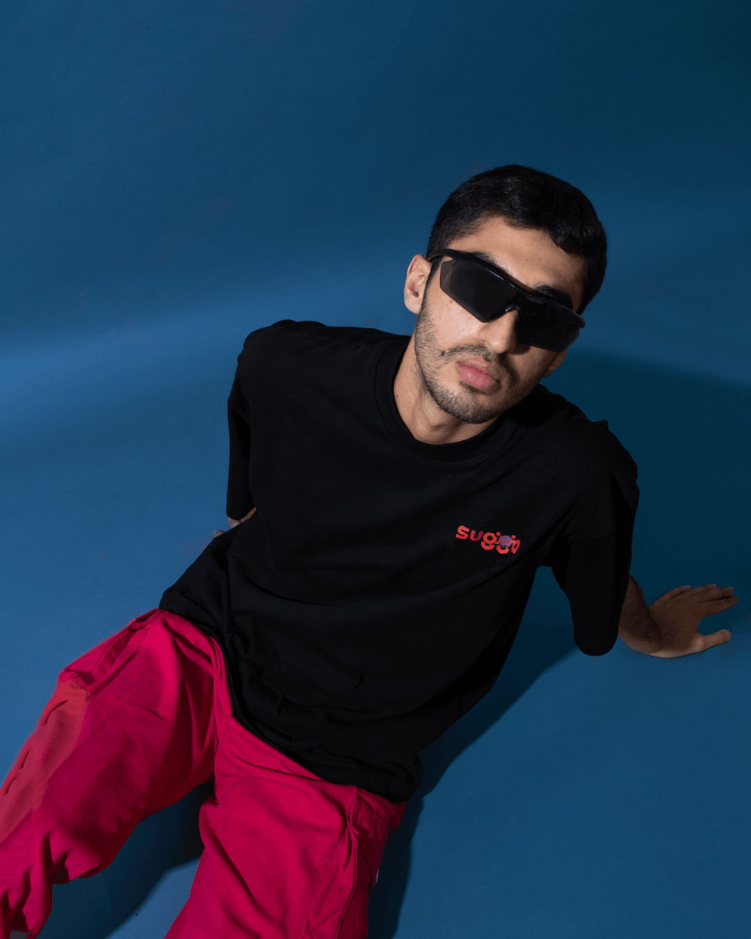 Man in black graphic t-shirt, red cargo pants, and black sunglasses on blue background