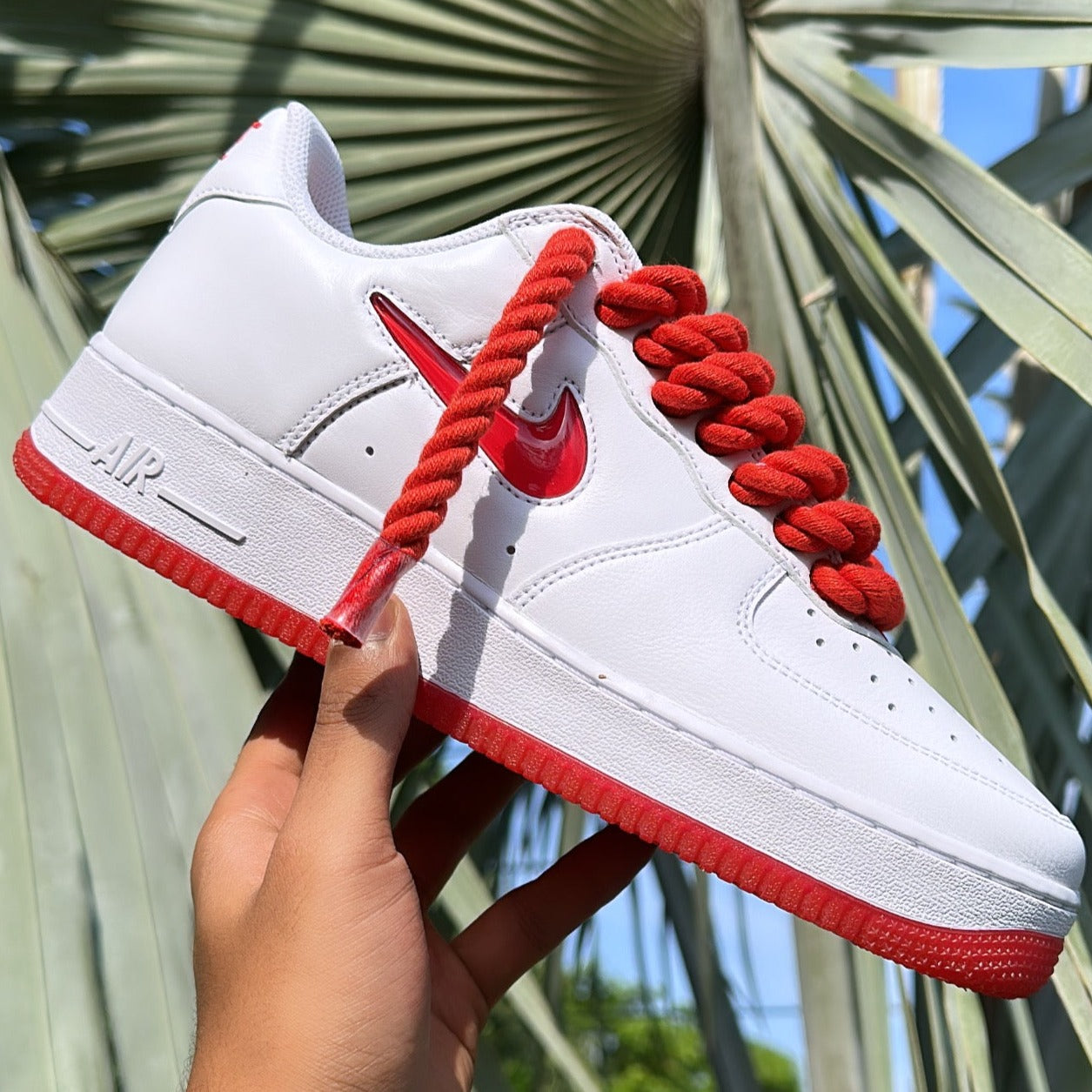 White and red Nike sneaker with thick red laces, held outdoors with palm leaves background