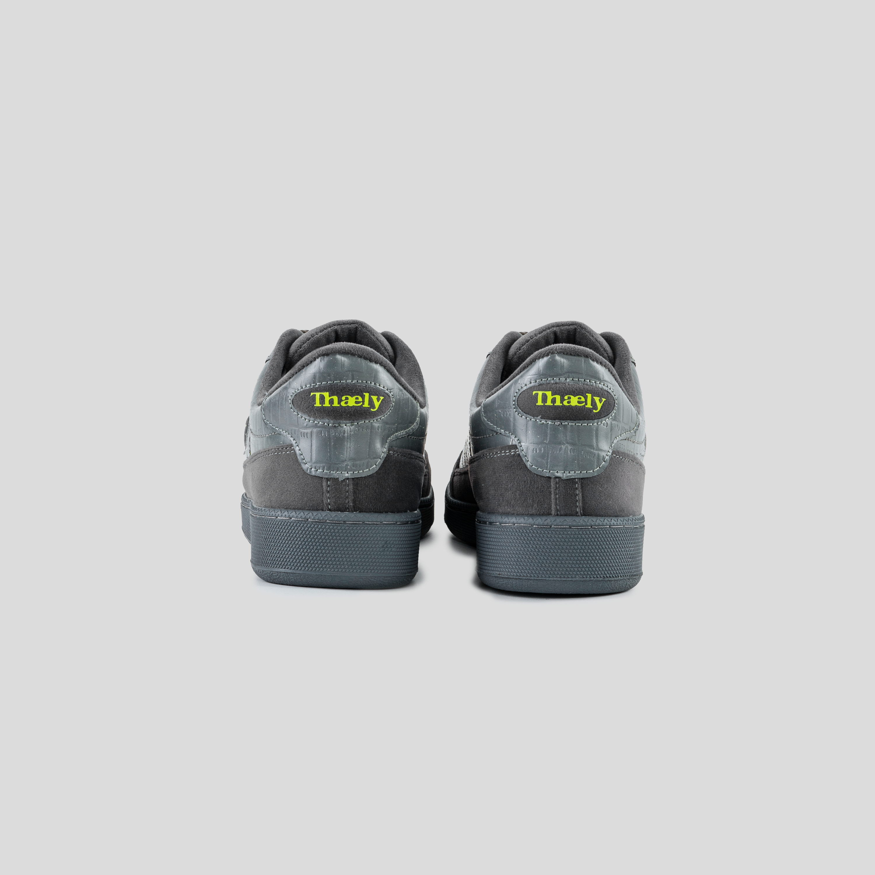 Back view of gray streetwear sneakers with suede details and neon yellow Thaely logo, on plain background