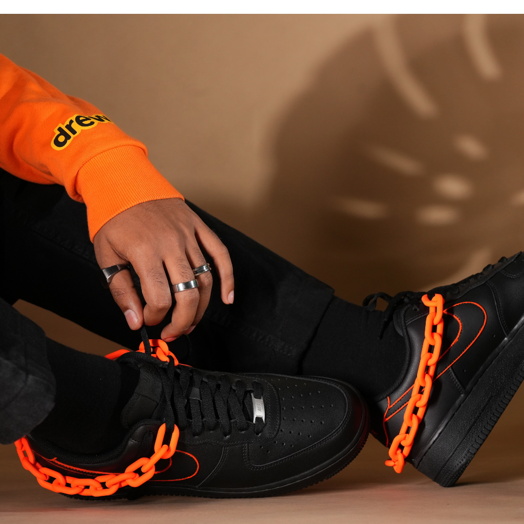 Black streetwear sneakers with orange chain and accents, worn with black pants and orange sleeve
