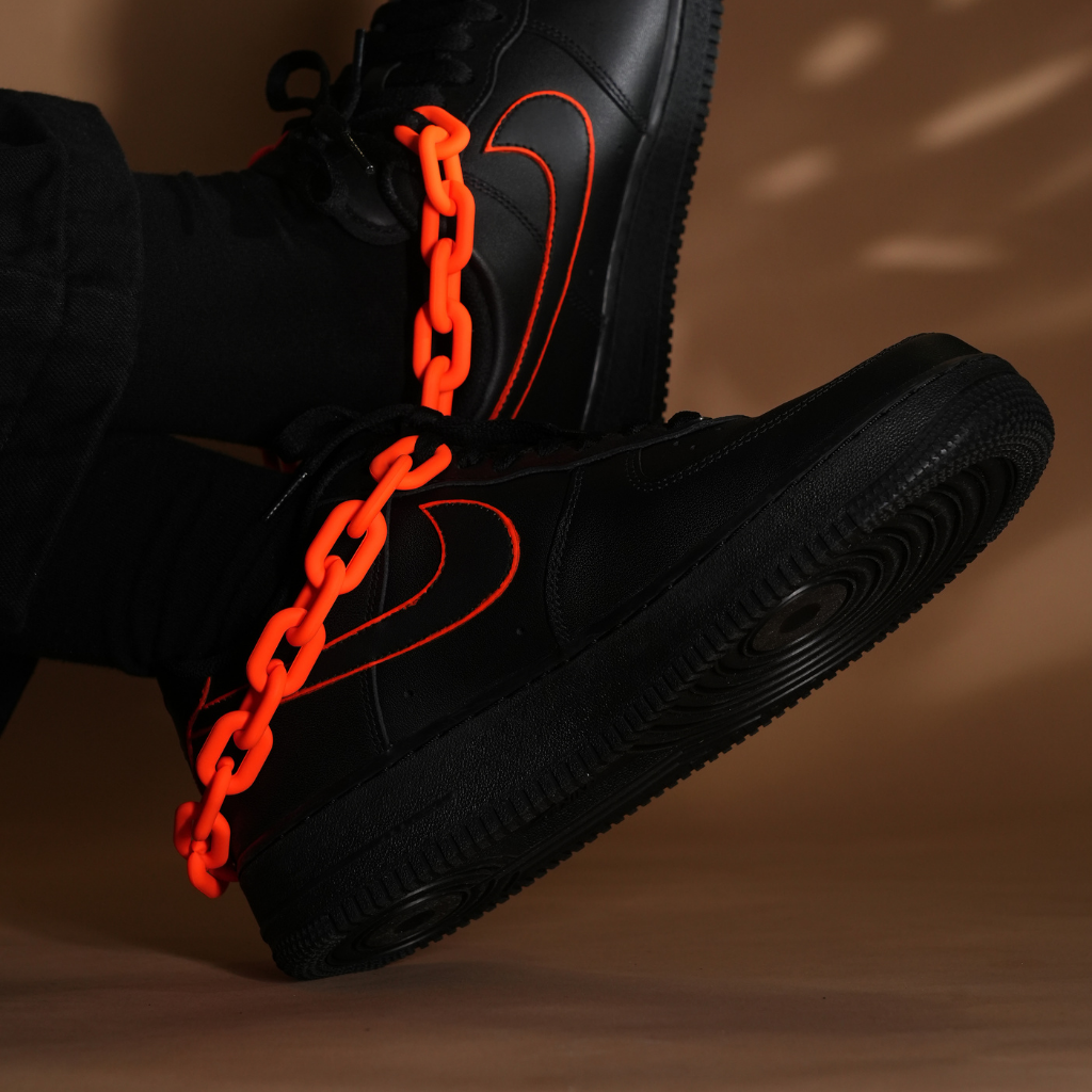 Black streetwear sneakers with neon orange outline and chunky orange chain on brown background