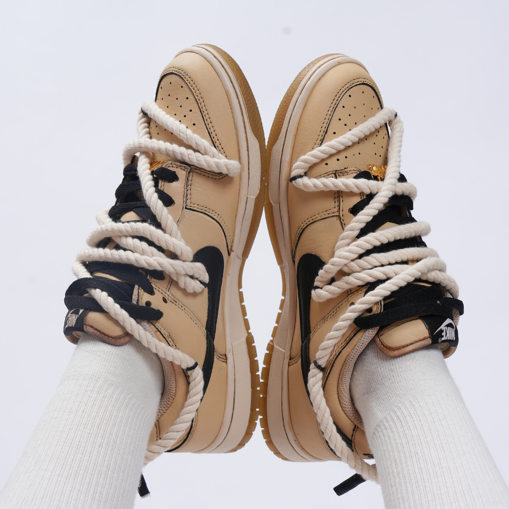 Beige and black Nike Dunk Low sneakers with thick rope laces on feet, white background