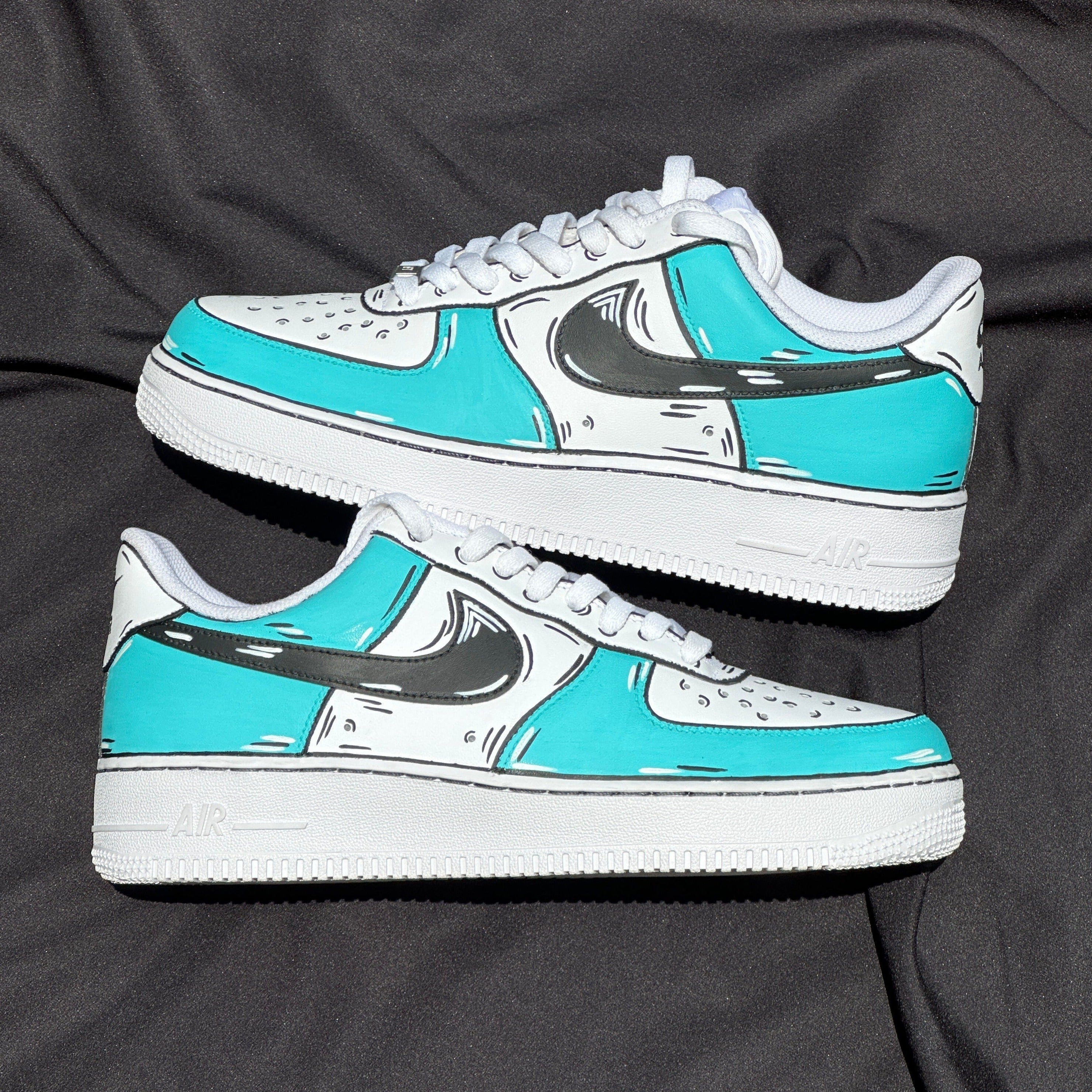 Custom teal and white sneakers with black graphic accents on a black fabric background.
