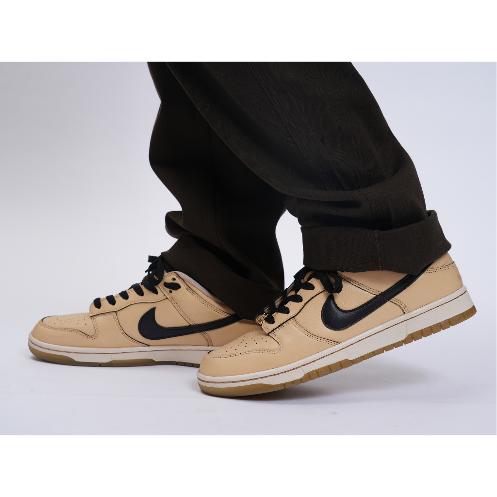 Tan and black Nike Dunk Low sneakers styled with black pants on a white background