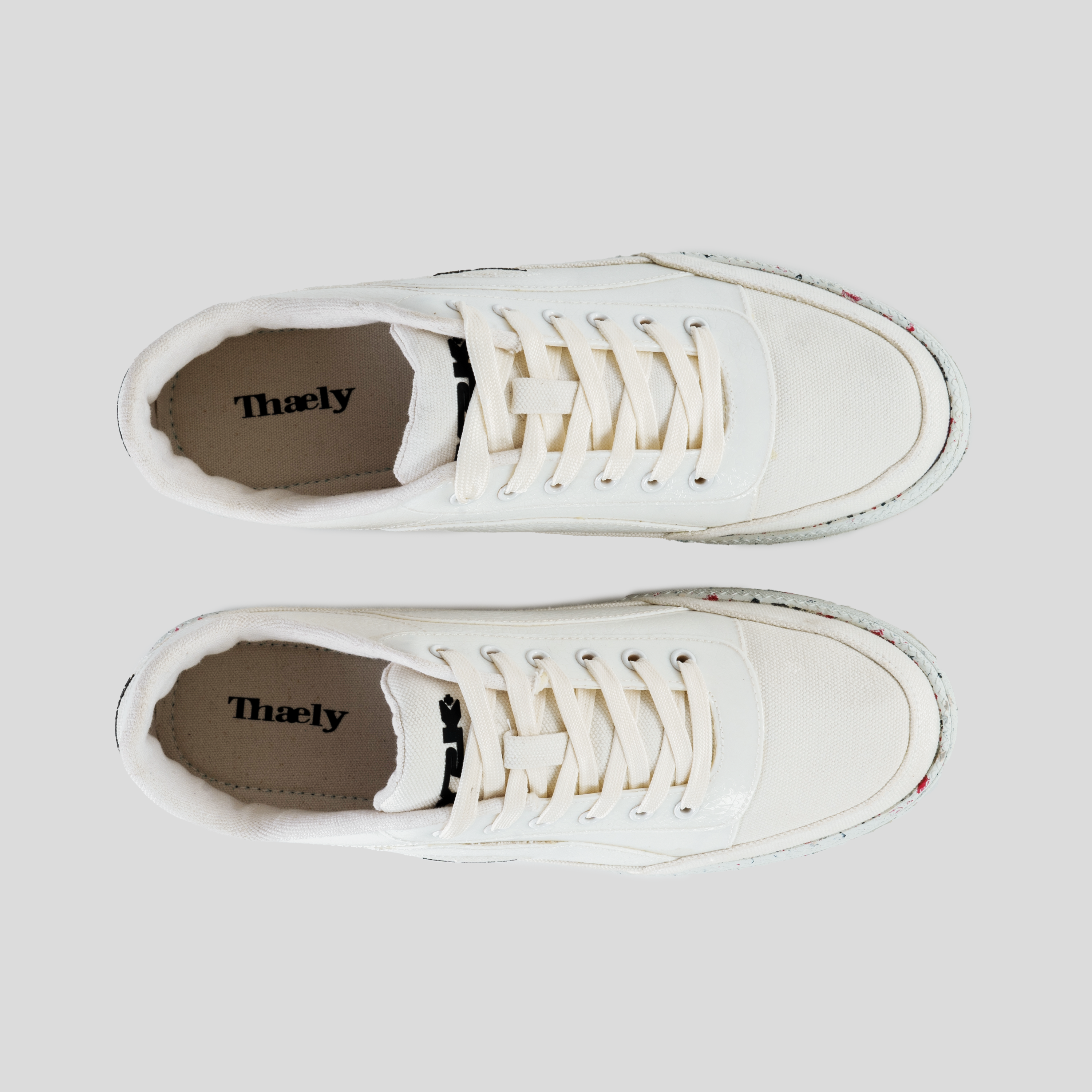 Top view of white low-top sneakers with canvas upper and recycled rubber soles, Thaely branding visible on insoles.