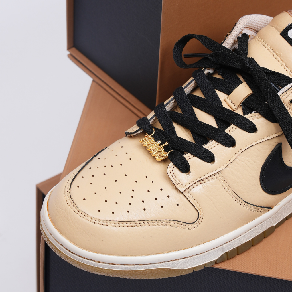 Beige leather sneaker with black laces, gold Nike lace tag, and black swoosh on stacked boxes