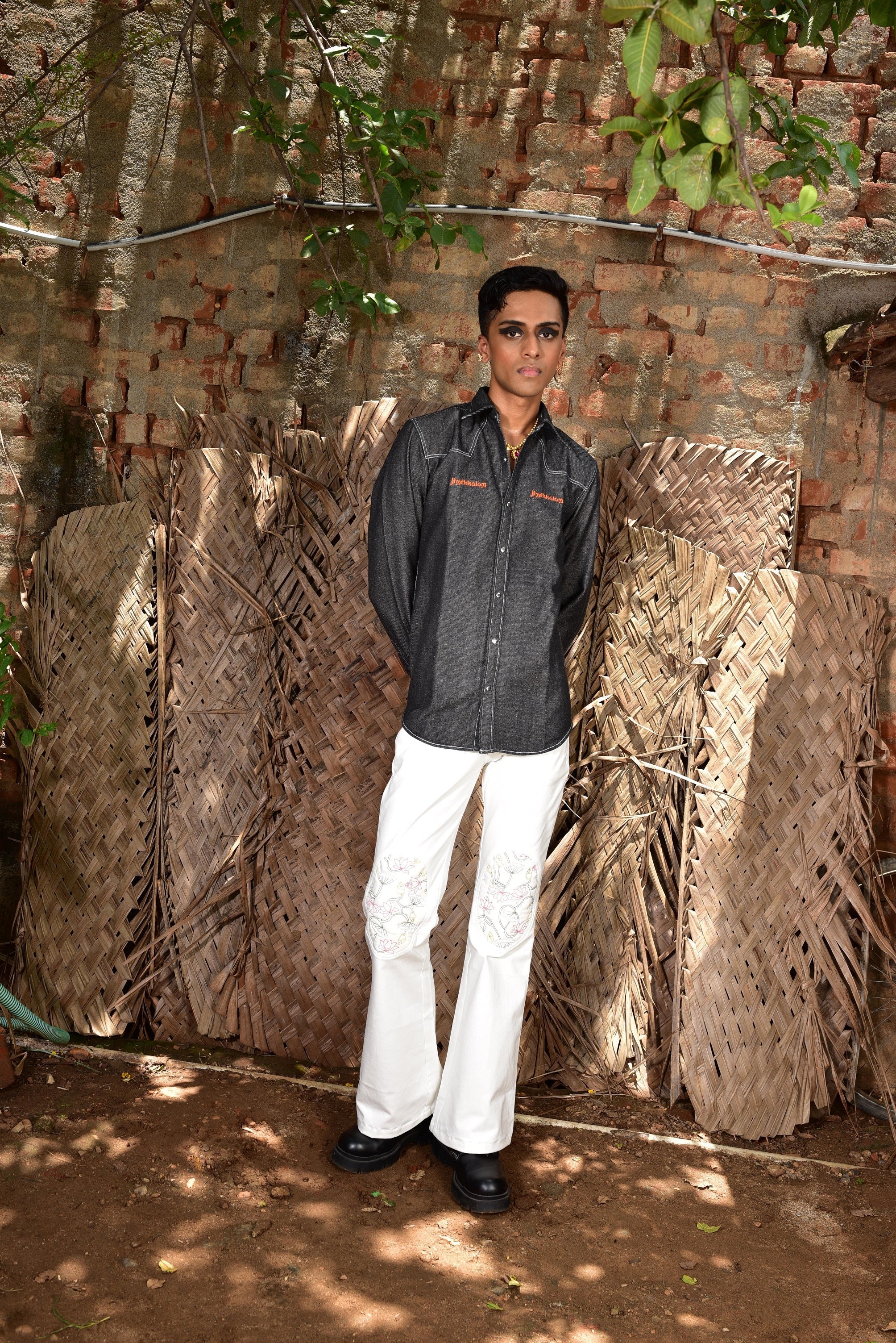 Male model wears black denim shirt and white embroidered pants, standing outdoors by rustic woven panels