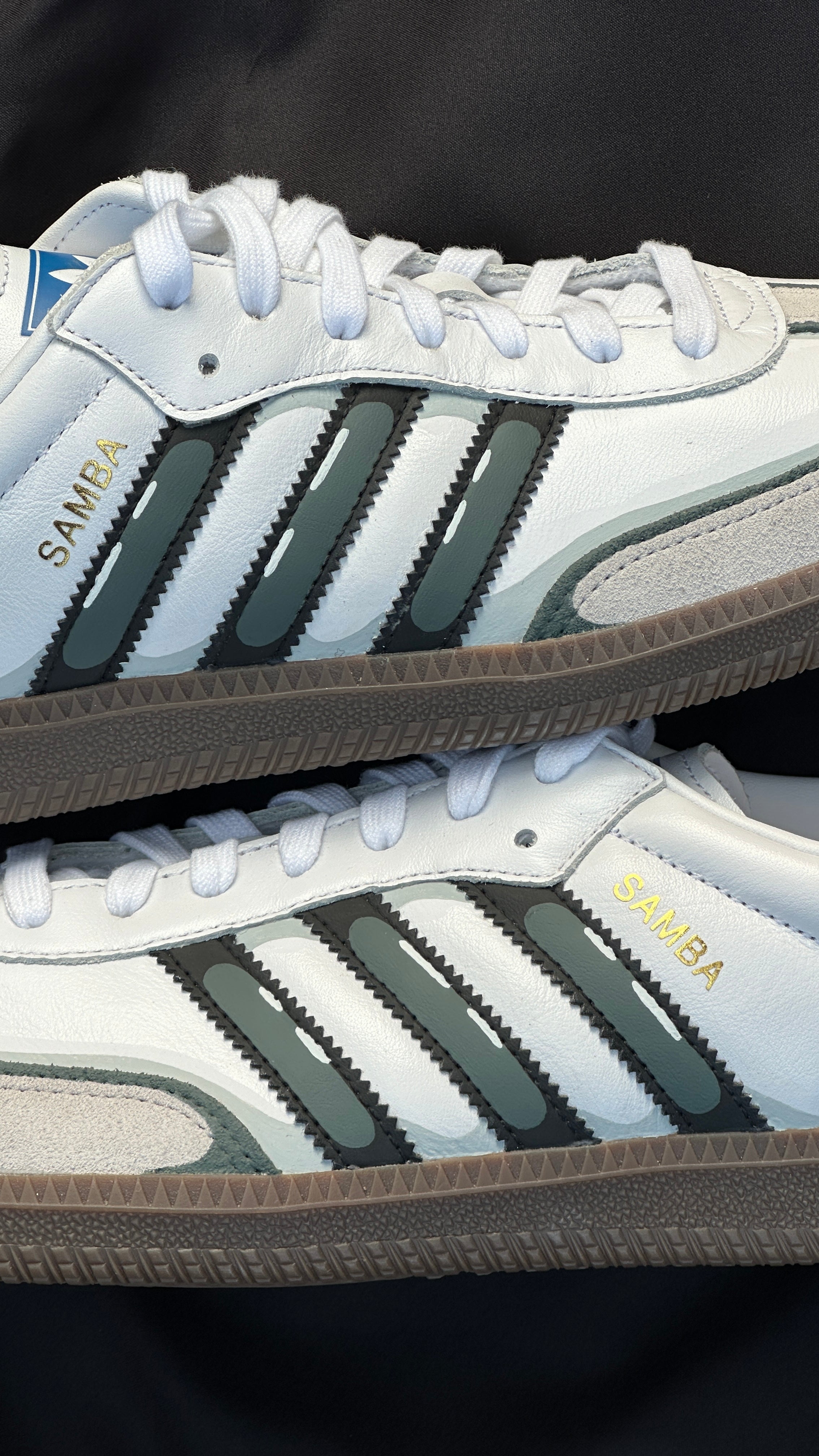 White Adidas Samba sneakers with black stripes, suede accents, and gum sole on dark background.