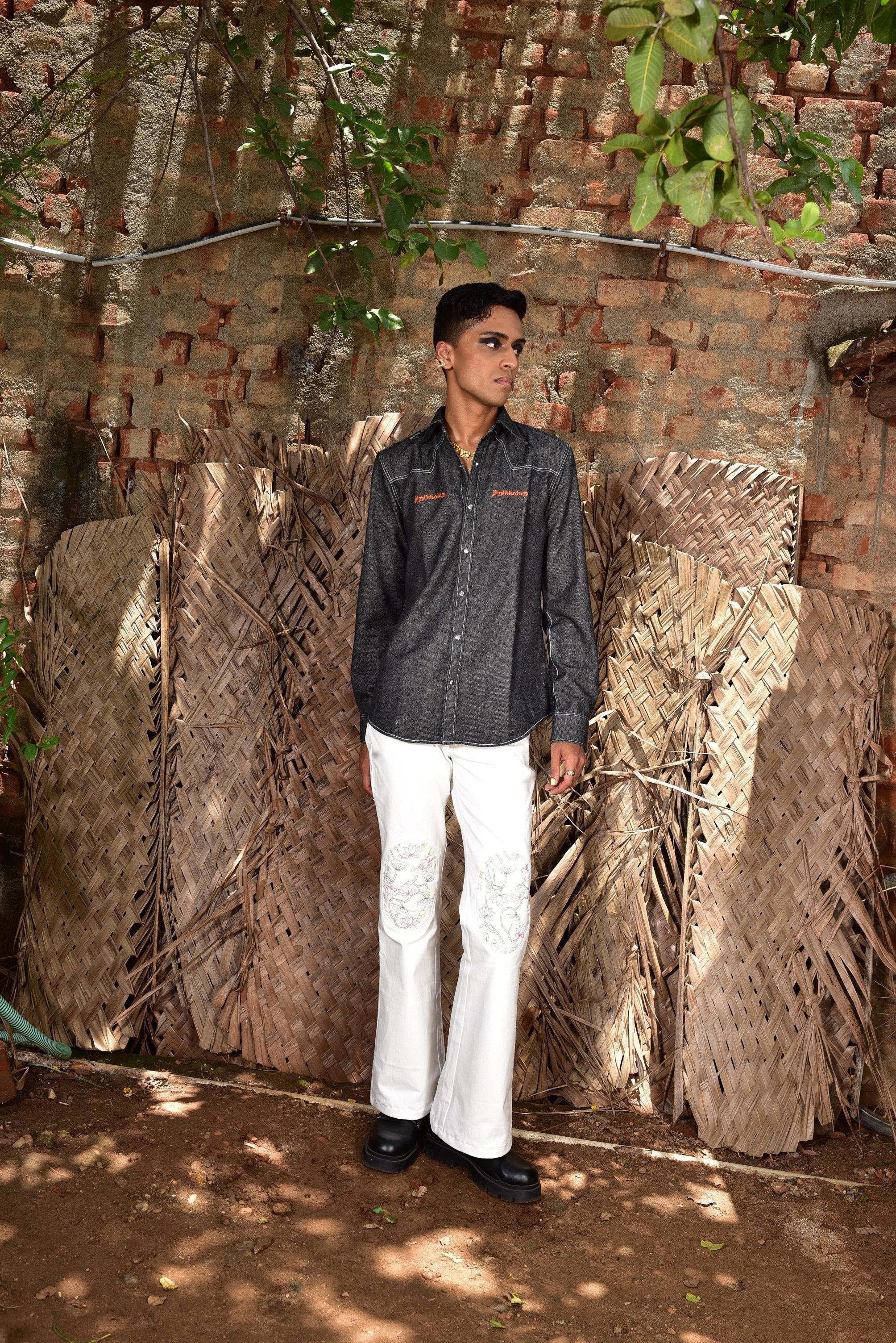Man in streetwear denim shirt and white embroidered pants poses outdoors by textured wall