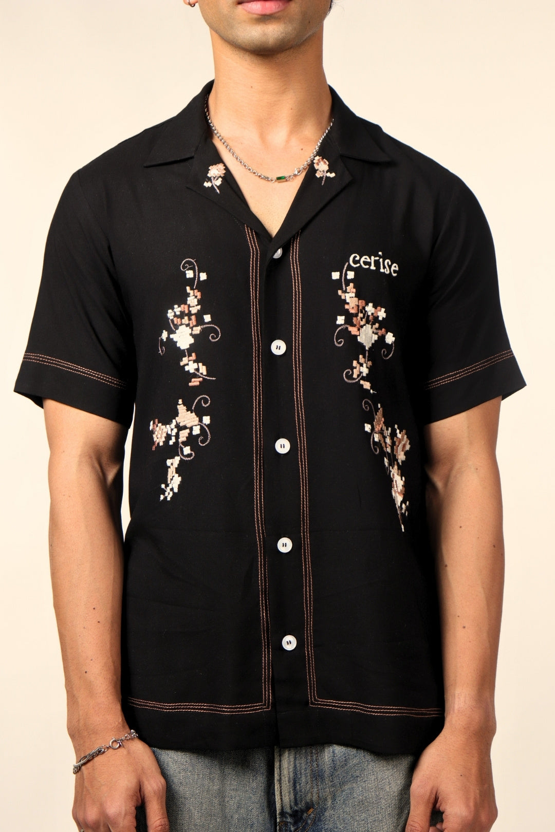 Man wearing a black short sleeve button-up shirt with floral embroidery and 'Cerise' text, paired with jeans.