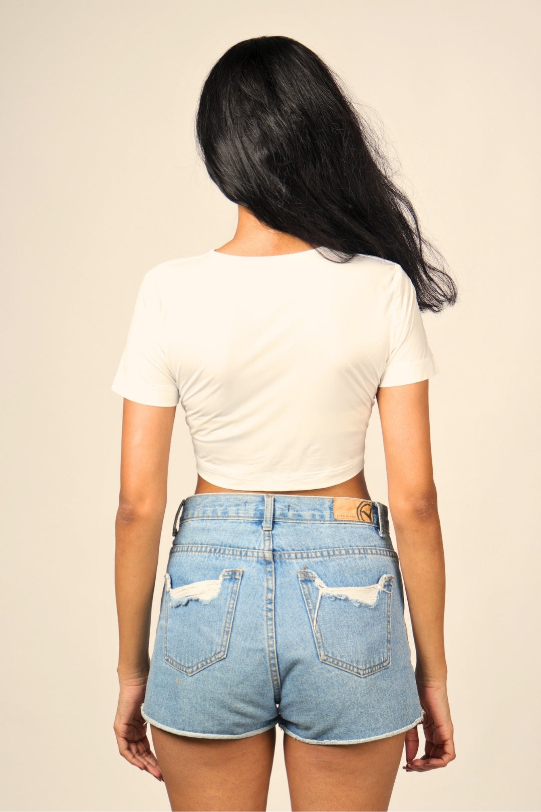 Woman wearing a white crop top and distressed high-waisted denim shorts, back view