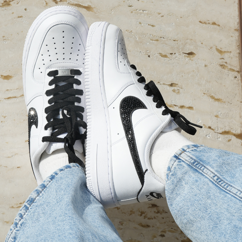 White sneakers with black sparkling swoosh, black laces, and blue jeans on stone surface