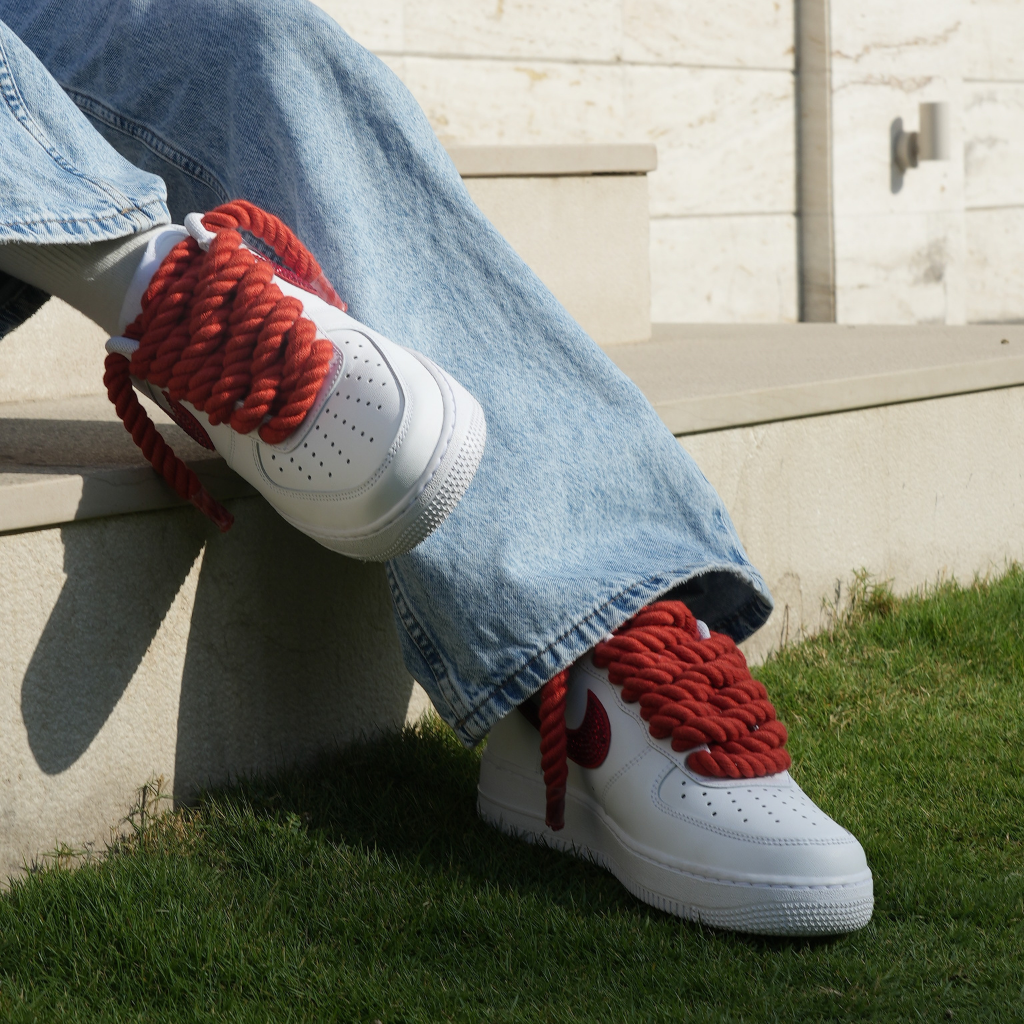 White streetwear sneakers with chunky red rope laces, worn with baggy jeans on outdoor steps