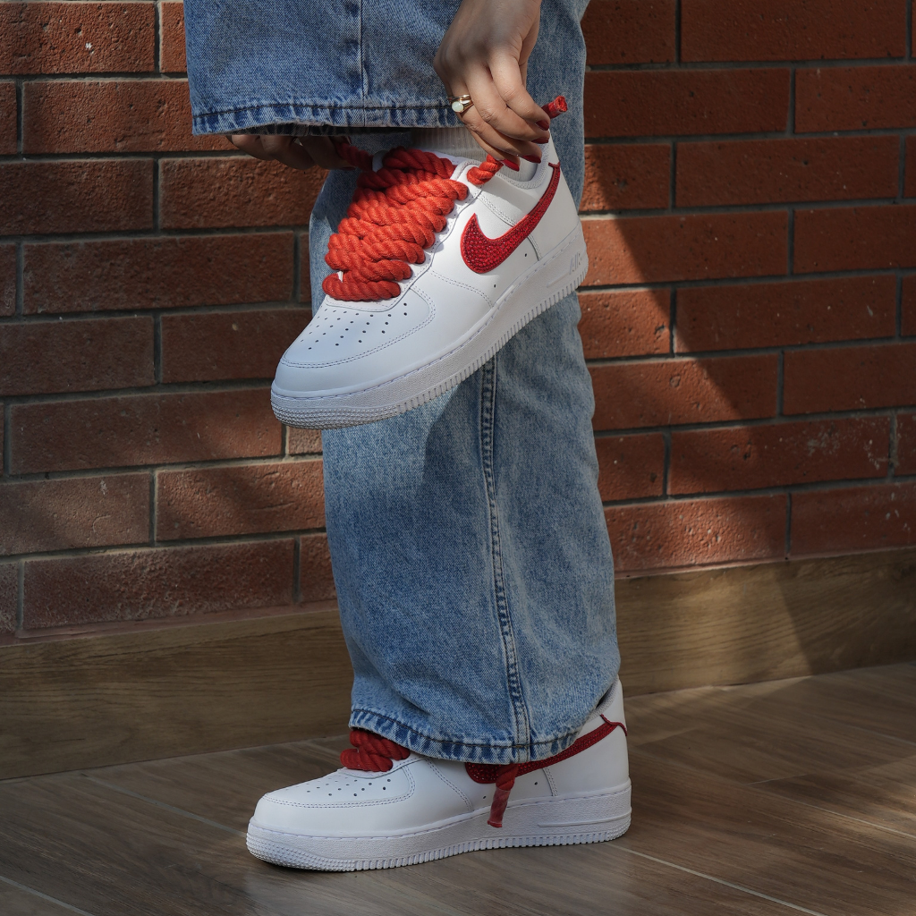 White Nike sneakers with red swoosh and thick red laces, styled with blue jeans.