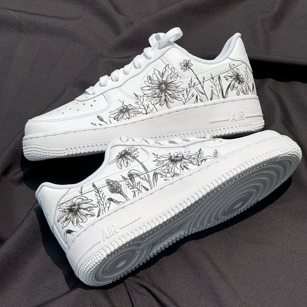 White streetwear sneakers with black floral design on side panels, displayed on black fabric