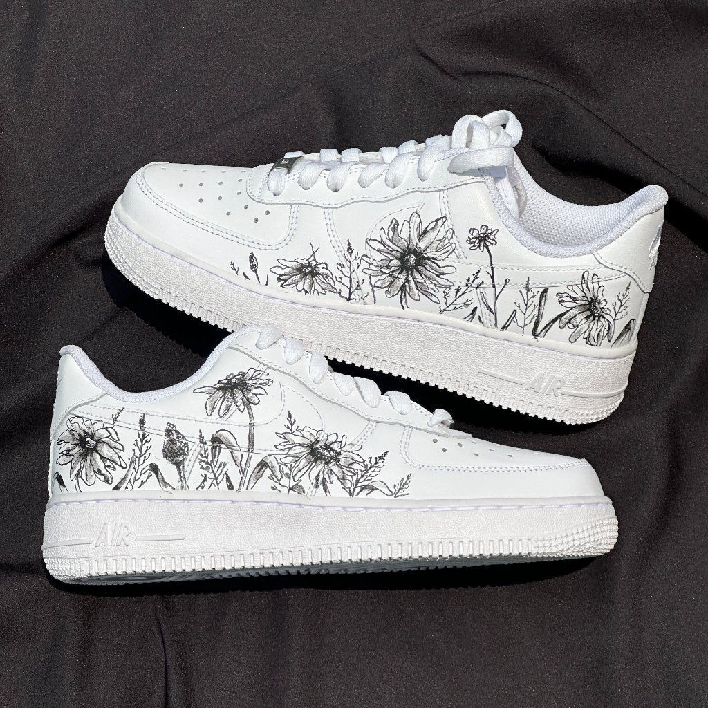 White sneakers with black floral illustrations on the sides, displayed on black fabric.