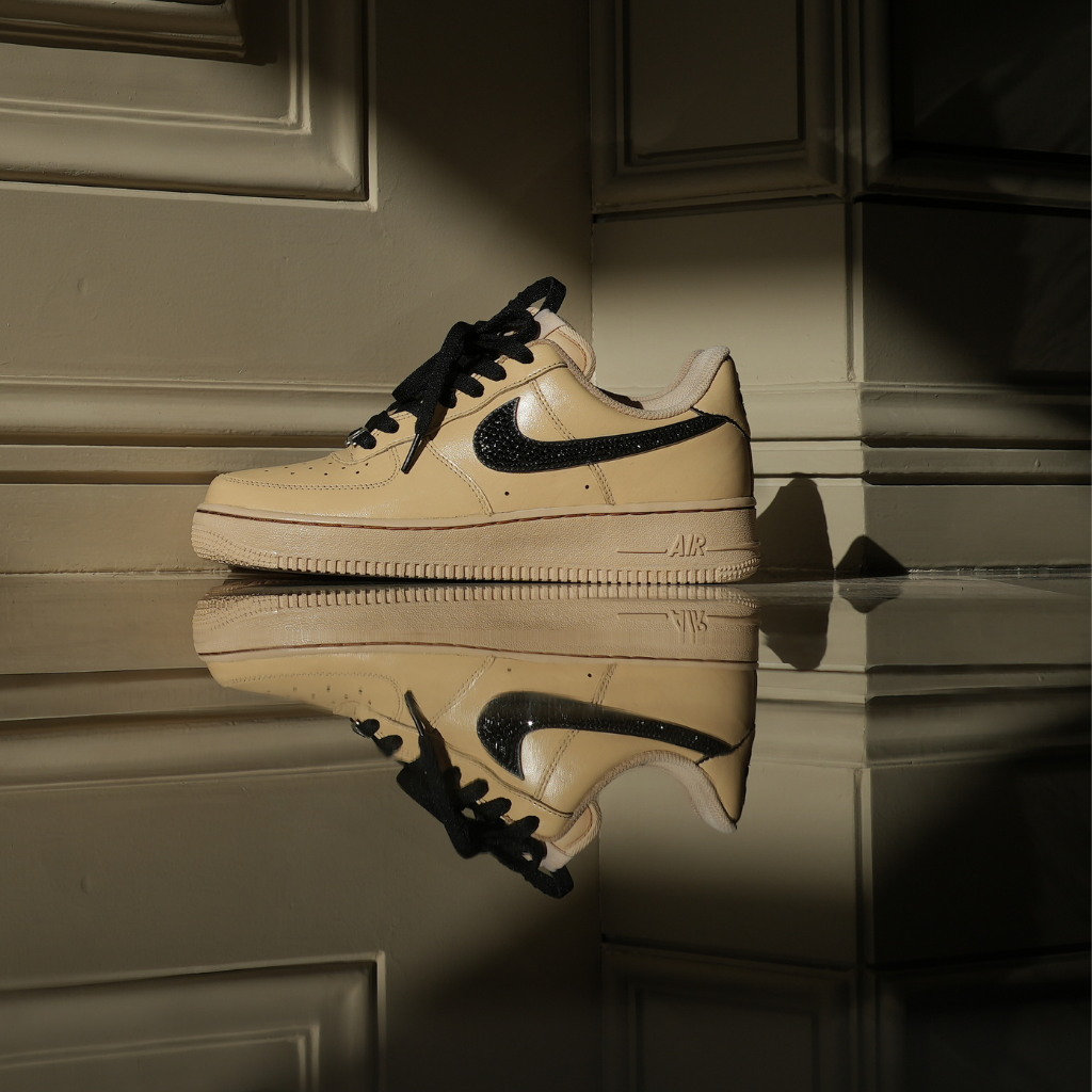 Beige Nike Air Force 1 sneaker with black swoosh and laces, reflected on glossy floor