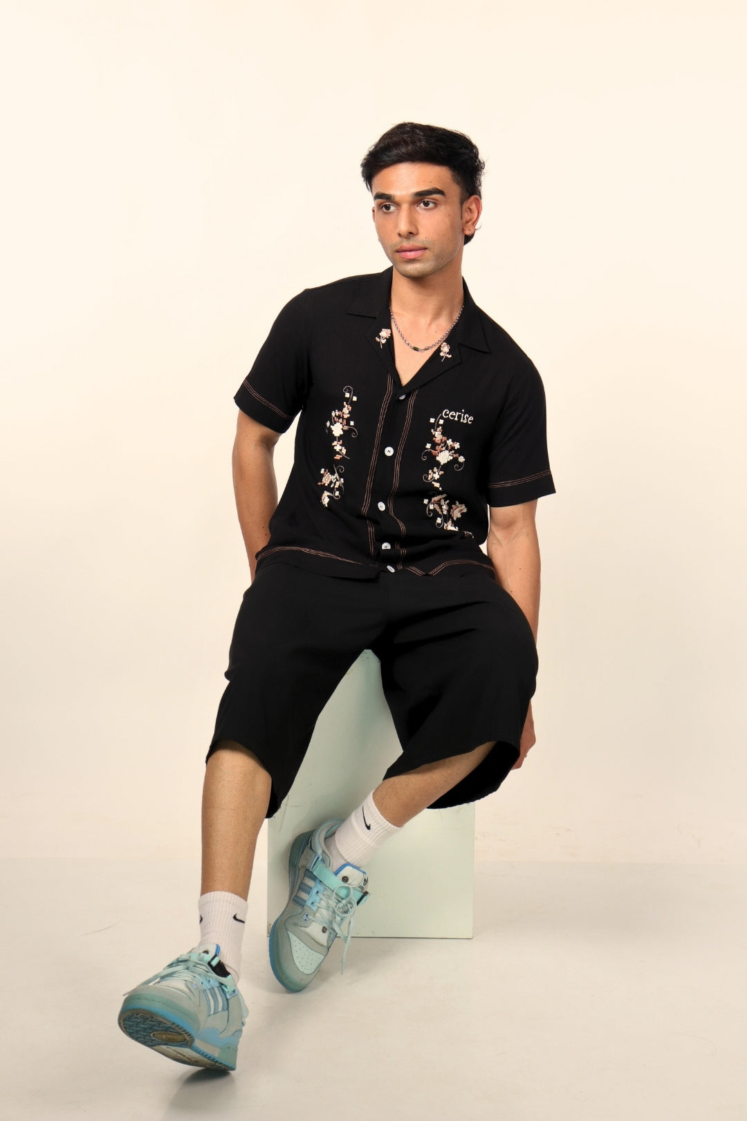 Man in black floral embroidered shirt, black shorts, and blue sneakers, streetwear style
