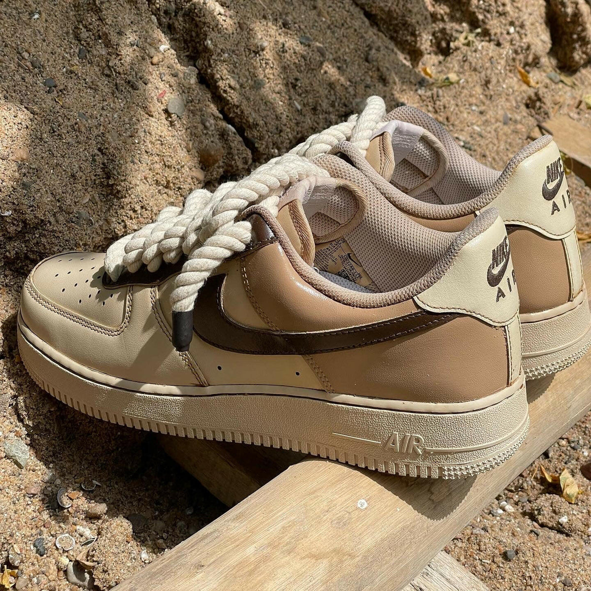 Beige and brown Nike Air Force 1 sneakers with rope laces on sand and wood outdoors
