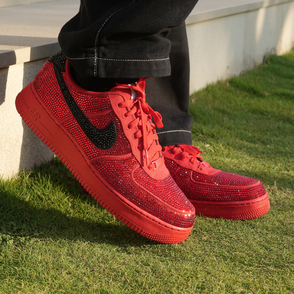 Red rhinestone-studded sneakers with black swoosh, streetwear style, worn outdoors on grass