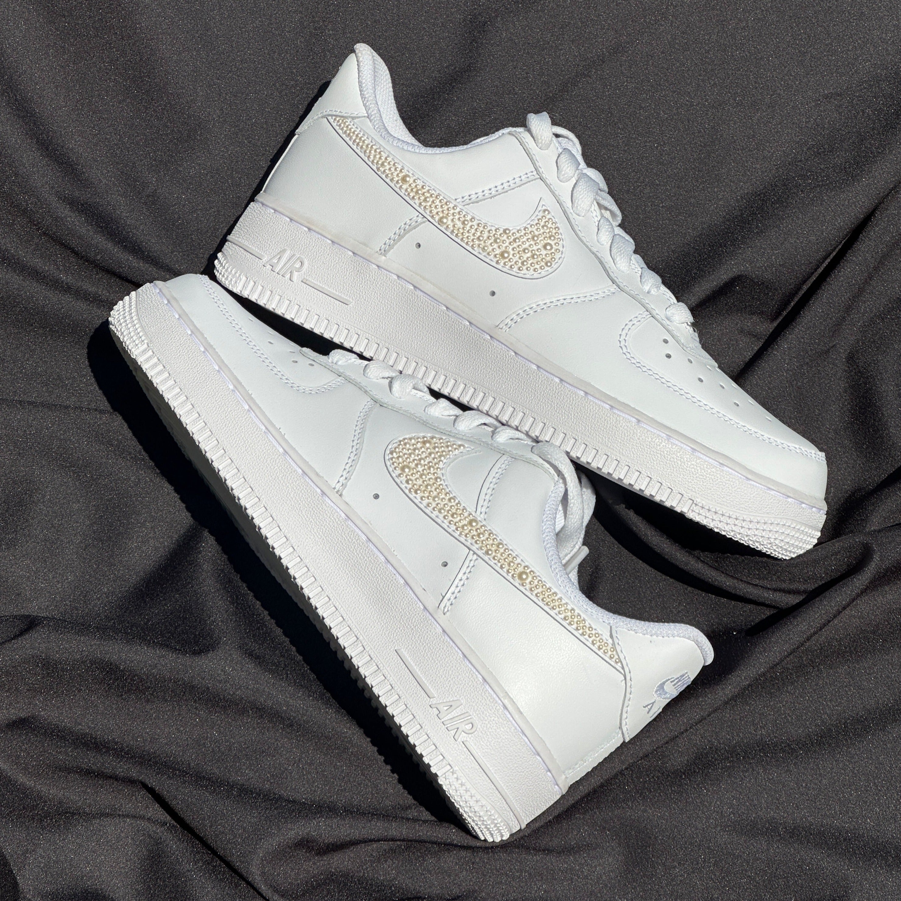 White designer sneakers with rhinestone swoosh detailing on black fabric background
