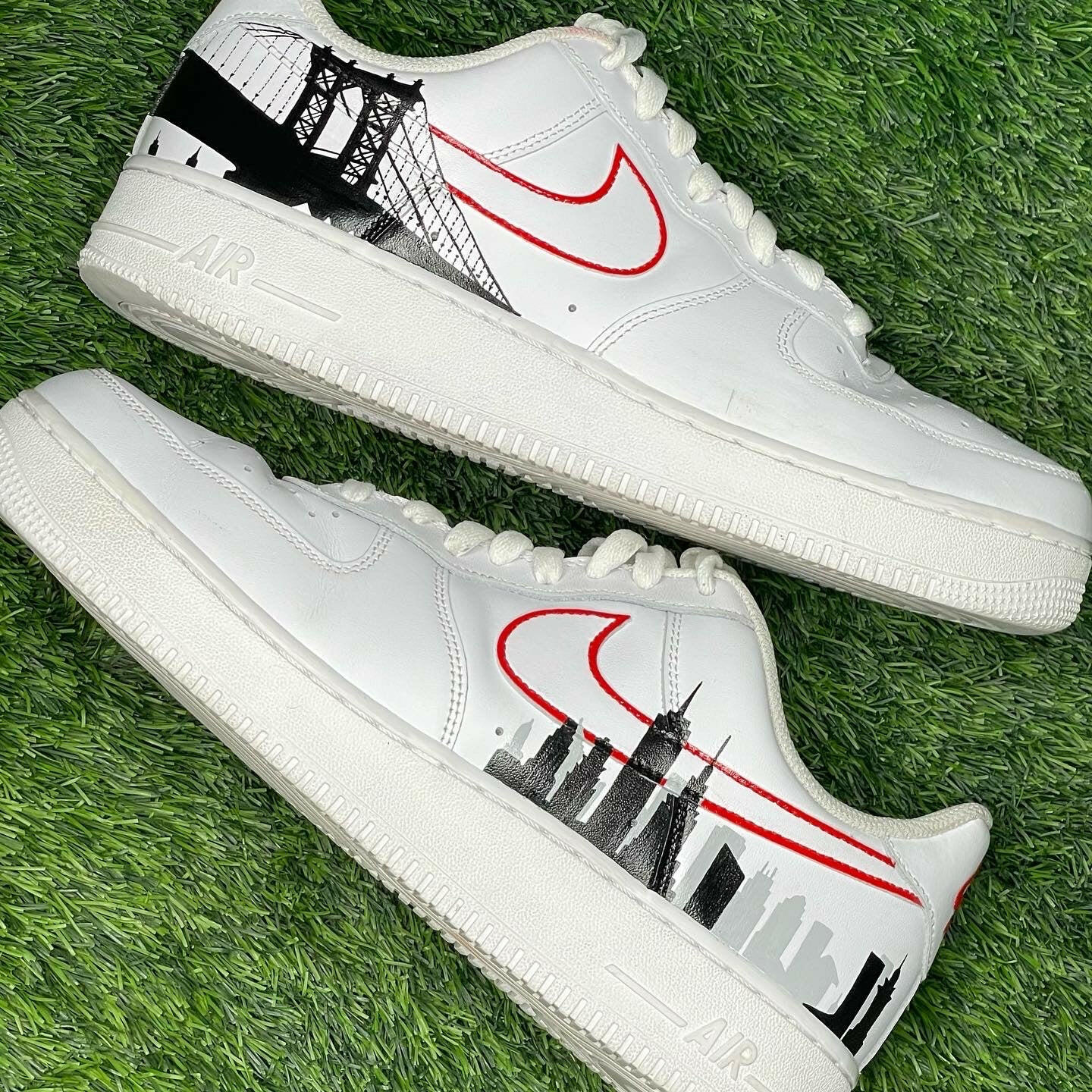 White Nike Air Force 1 sneakers with red Swoosh and black city skyline, bridge graphic on grass