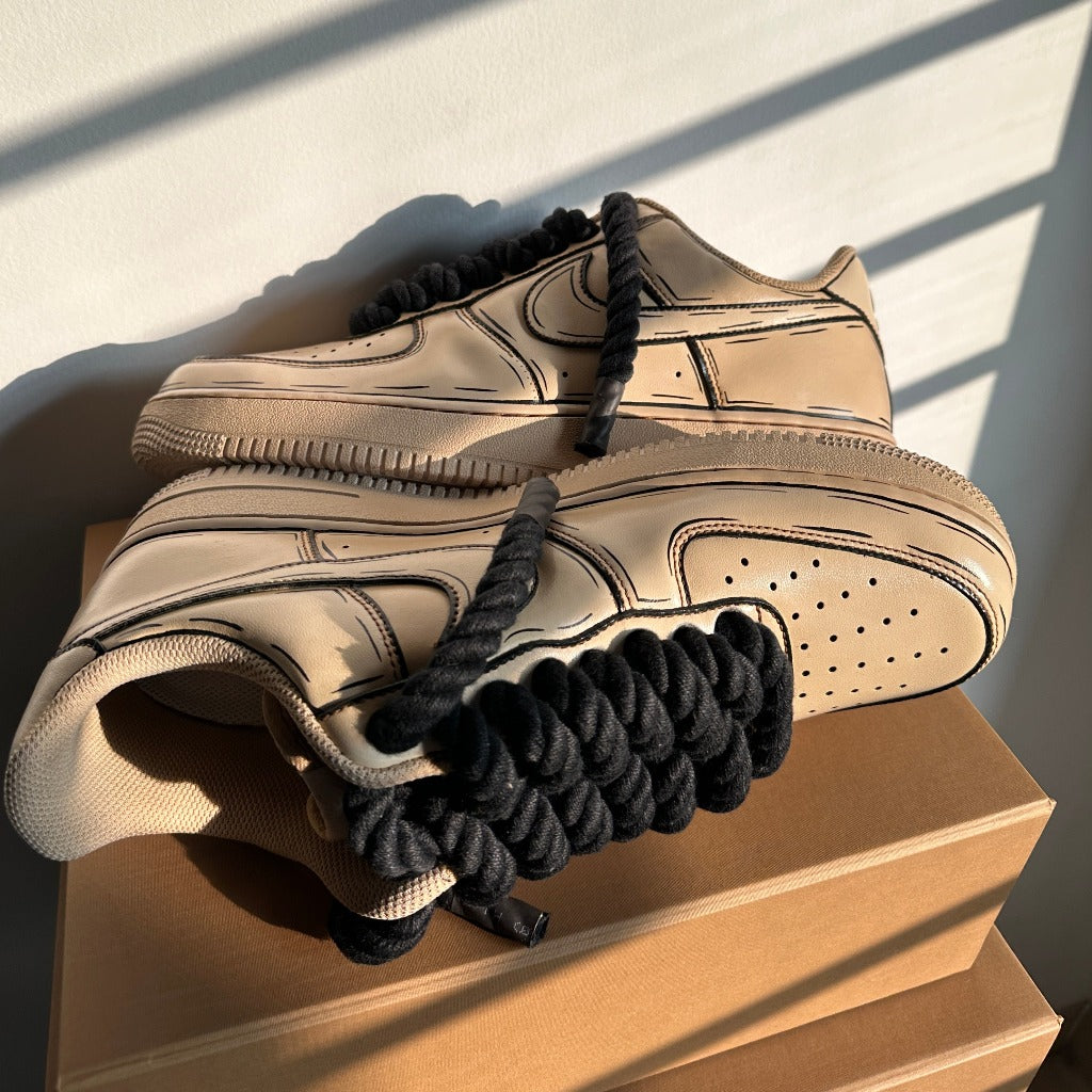 Beige streetwear sneakers with bold black laces and cartoon-style outlines on a box