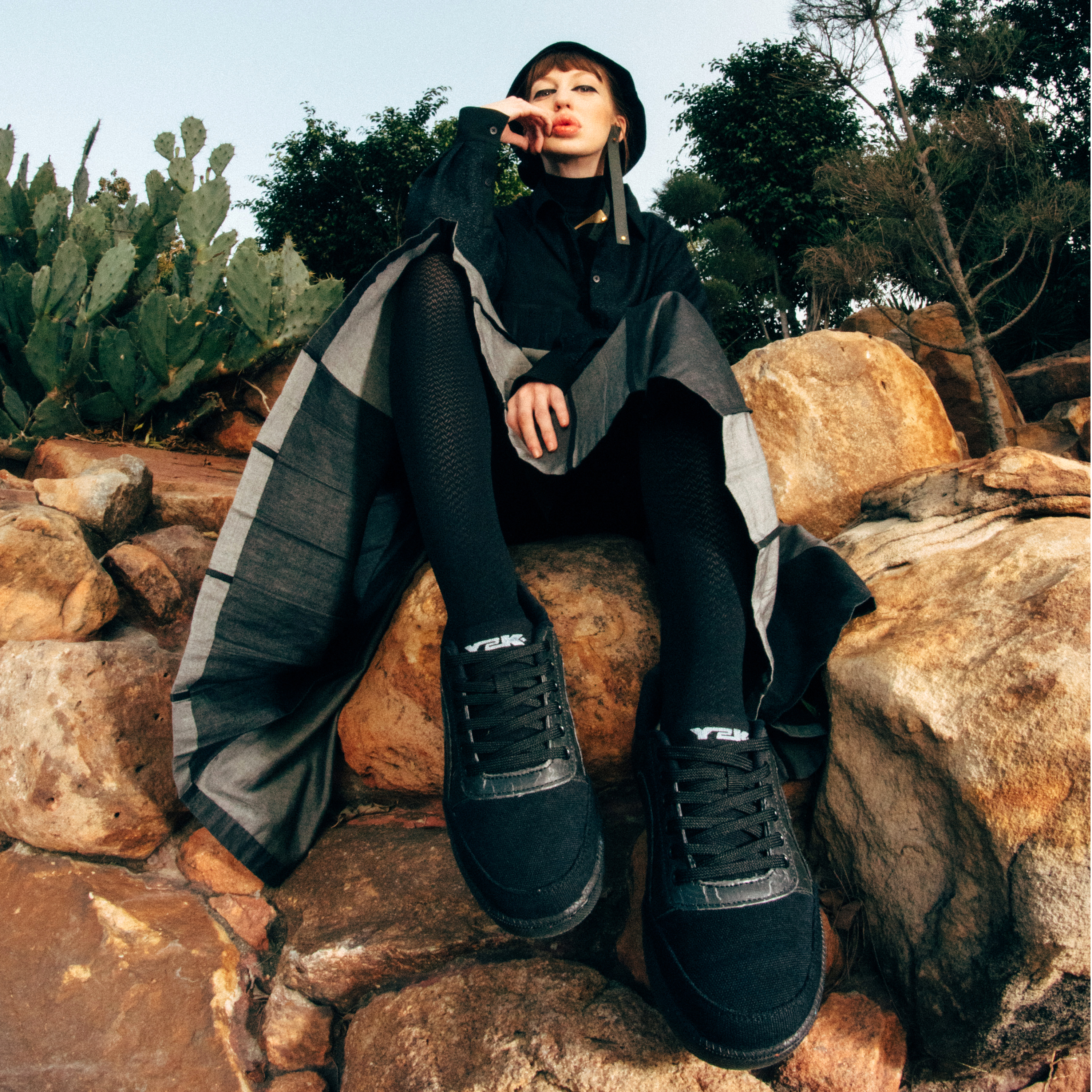 Woman in black streetwear and bucket hat sitting on rocks, wearing black sneakers outdoors