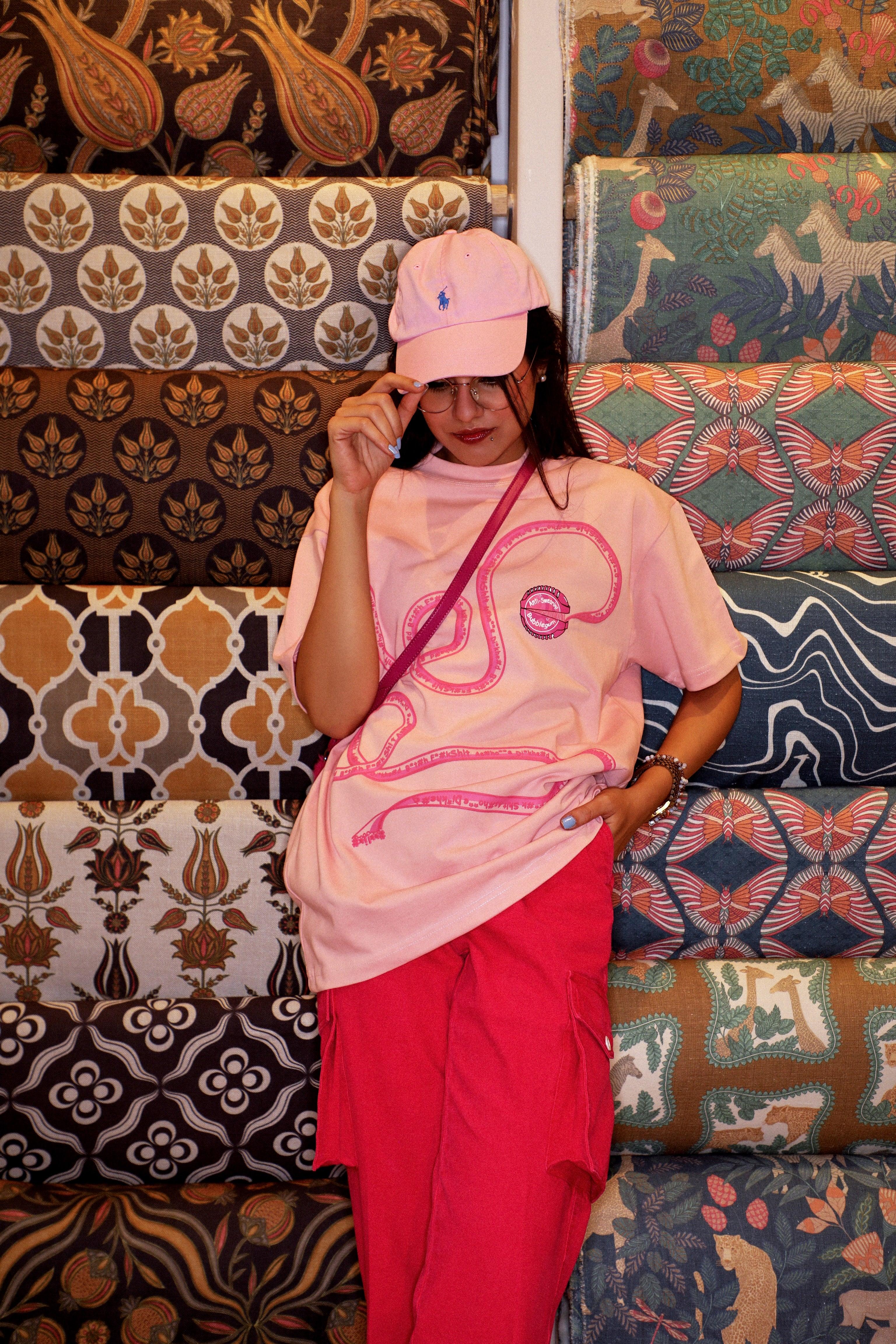 Woman in pink graphic streetwear t-shirt, pink cap, and red pants posing by colorful patterned fabrics.