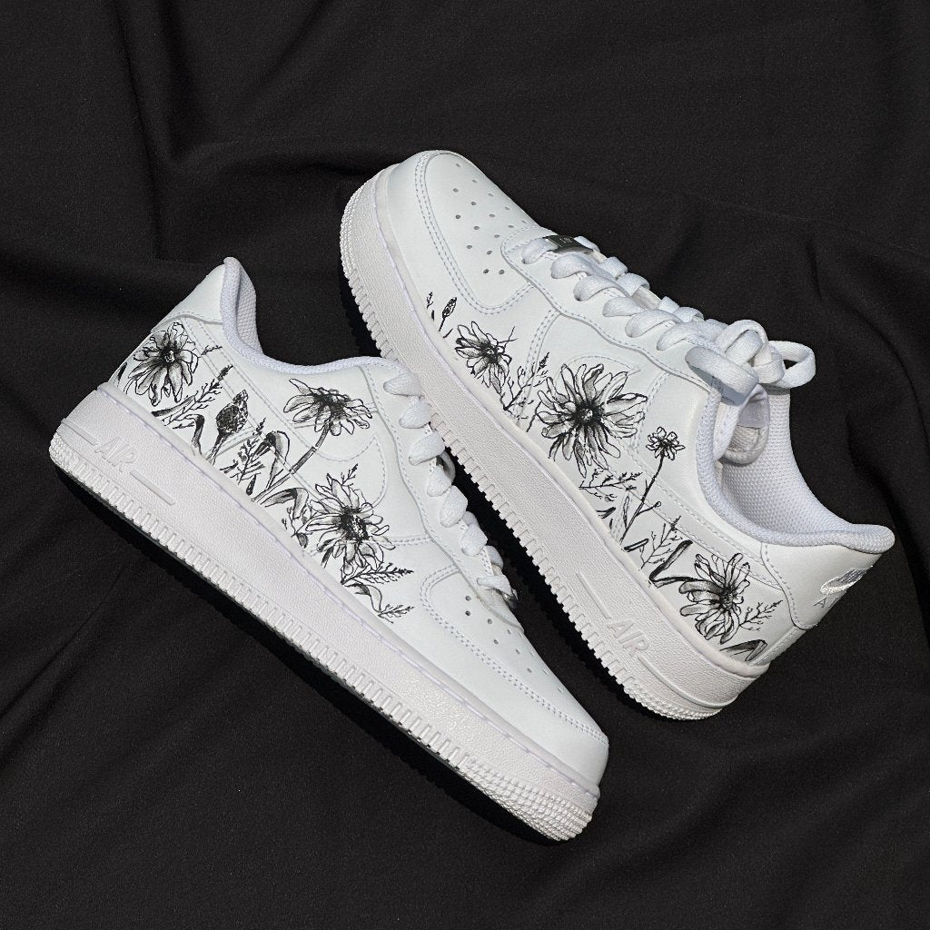 White streetwear sneakers with black floral illustrations on the sides, displayed on black fabric.