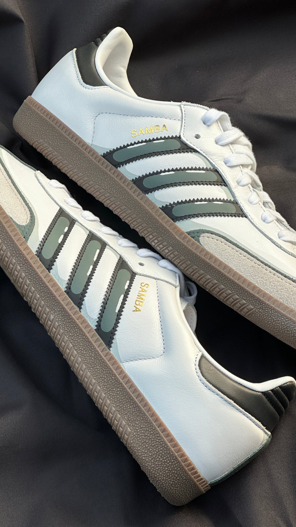 White Adidas Samba sneakers with black stripes and gum sole on a dark fabric background