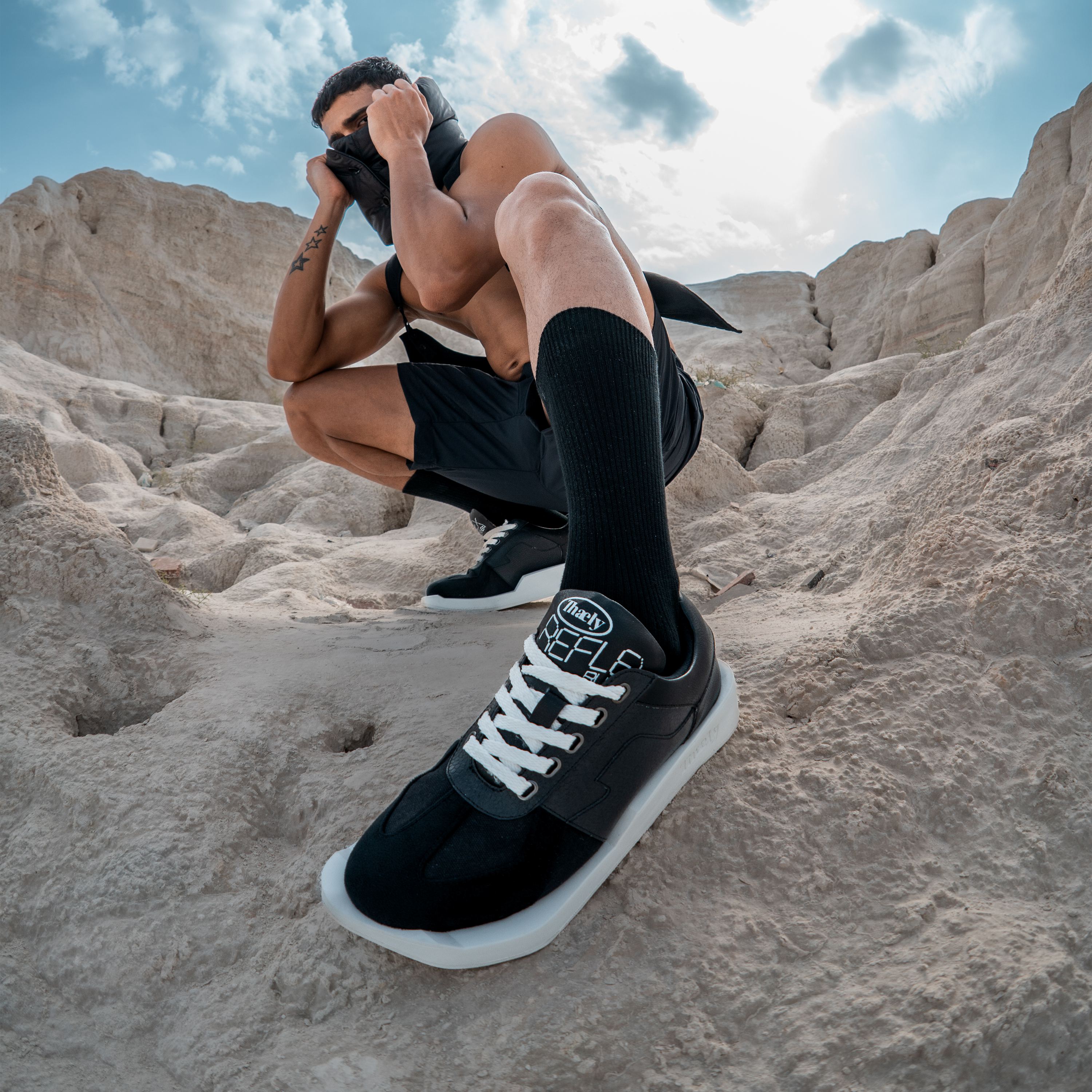 Man posing outdoors on rocky terrain wearing black streetwear sneakers and black athletic shorts.