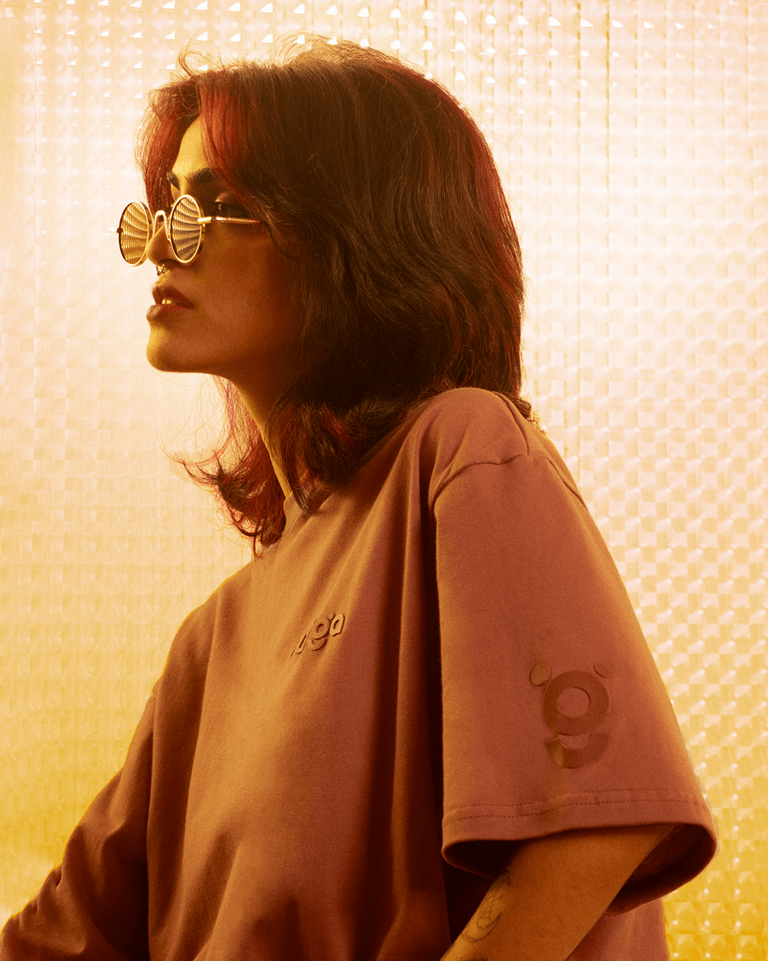 Person wearing oversized brown streetwear t-shirt and round sunglasses, warm light background