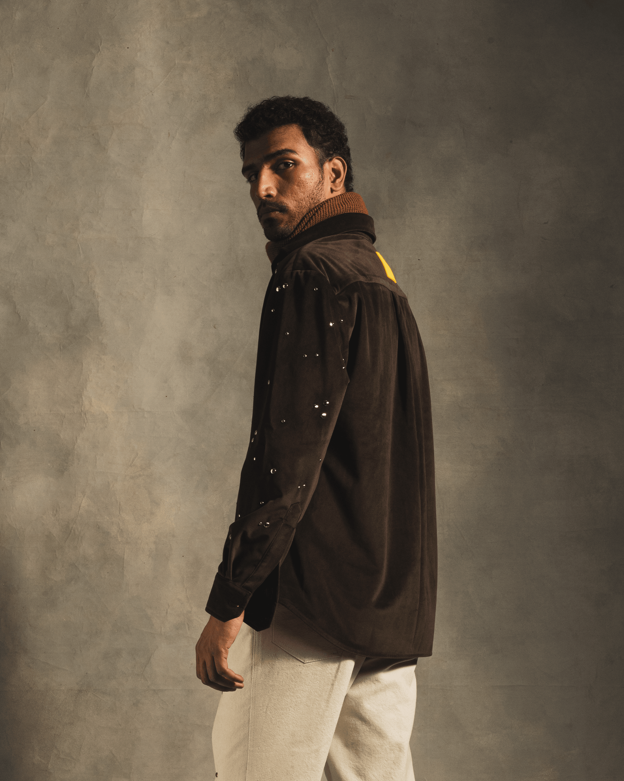 Man wearing brown oversized shirt with metallic rivet details and white pants, streetwear style.