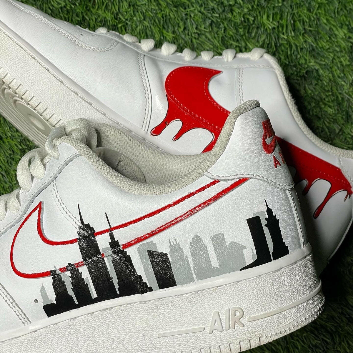 White streetwear sneakers with red Swoosh, skyline graphic, and drip detail on green grass