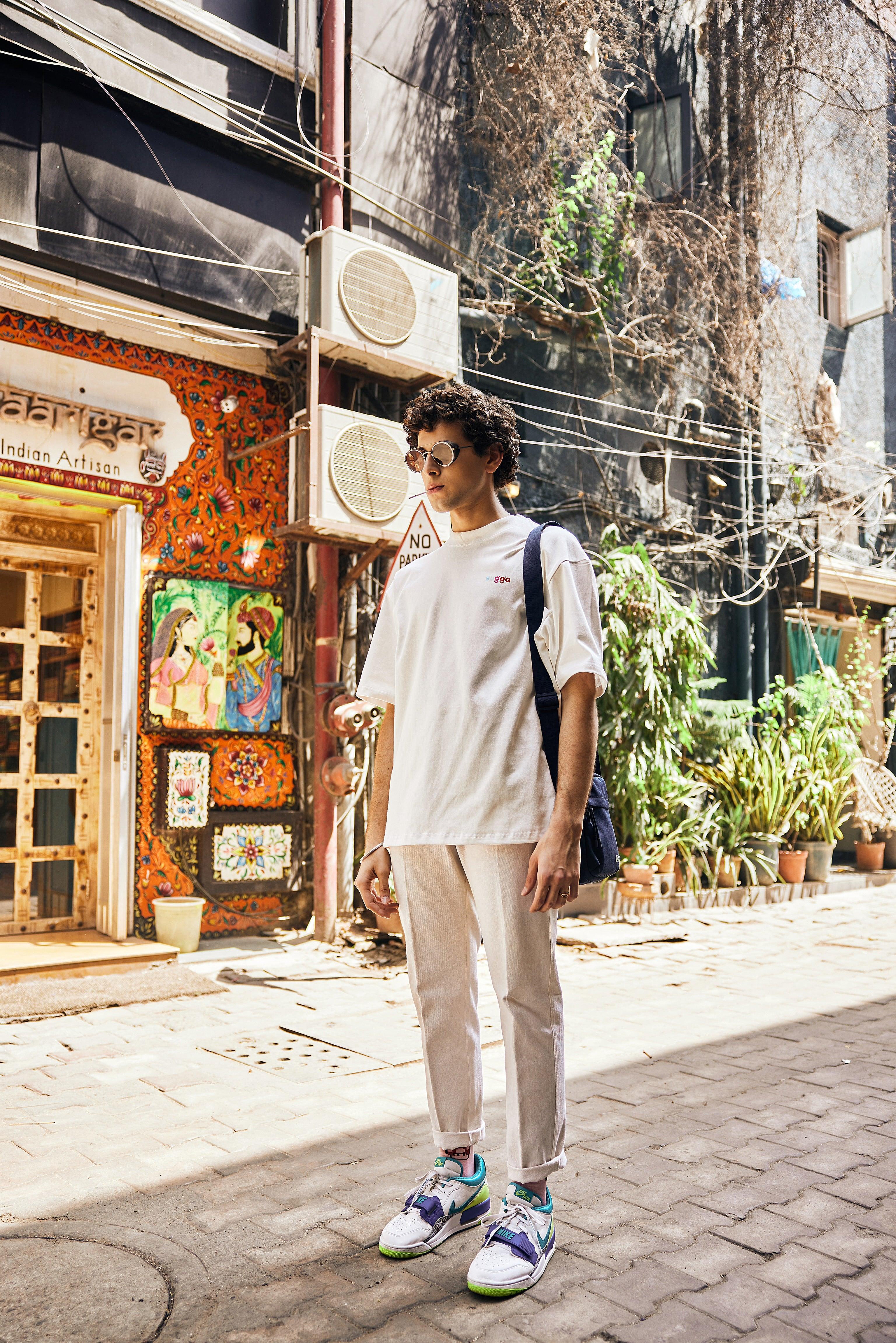 Man wearing streetwear outfit with white oversized t-shirt, beige pants, and colorful sneakers standing in an urban alleyway.