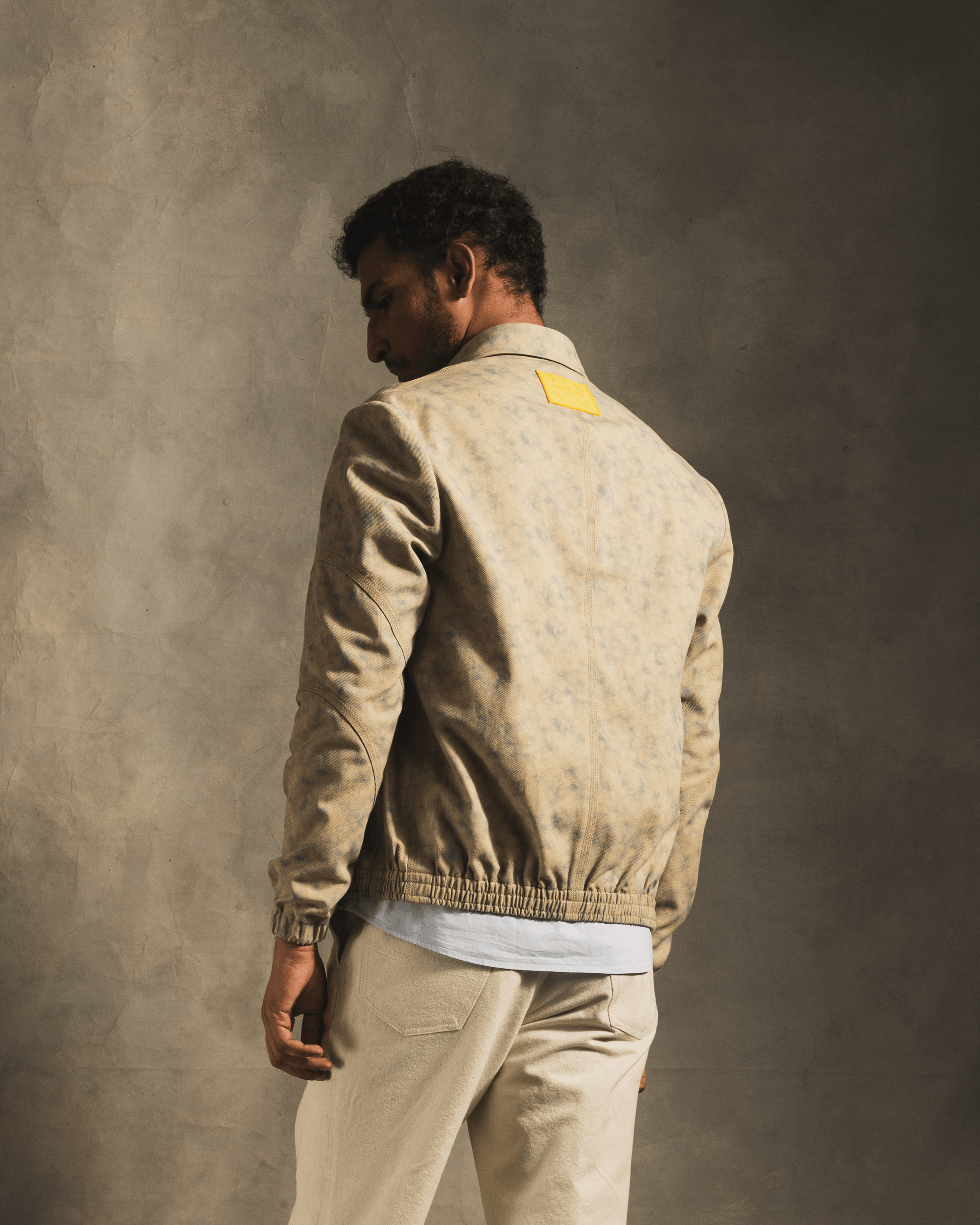 Man wearing a beige, cloud-pattern streetwear jacket with yellow patch, neutral background