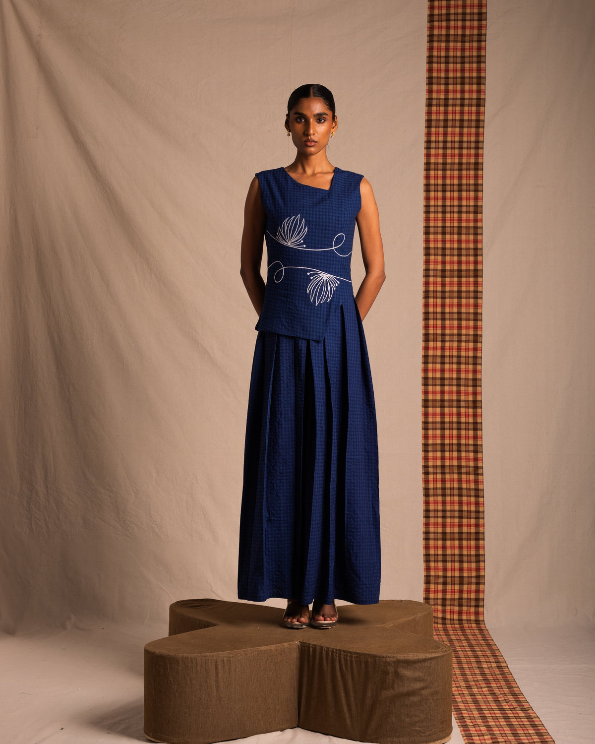 Woman in a navy blue sleeveless dress with white embroidery, standing on a brown pedestal in a studio with neutral and plaid fabric backdrop, modern fashion photography.