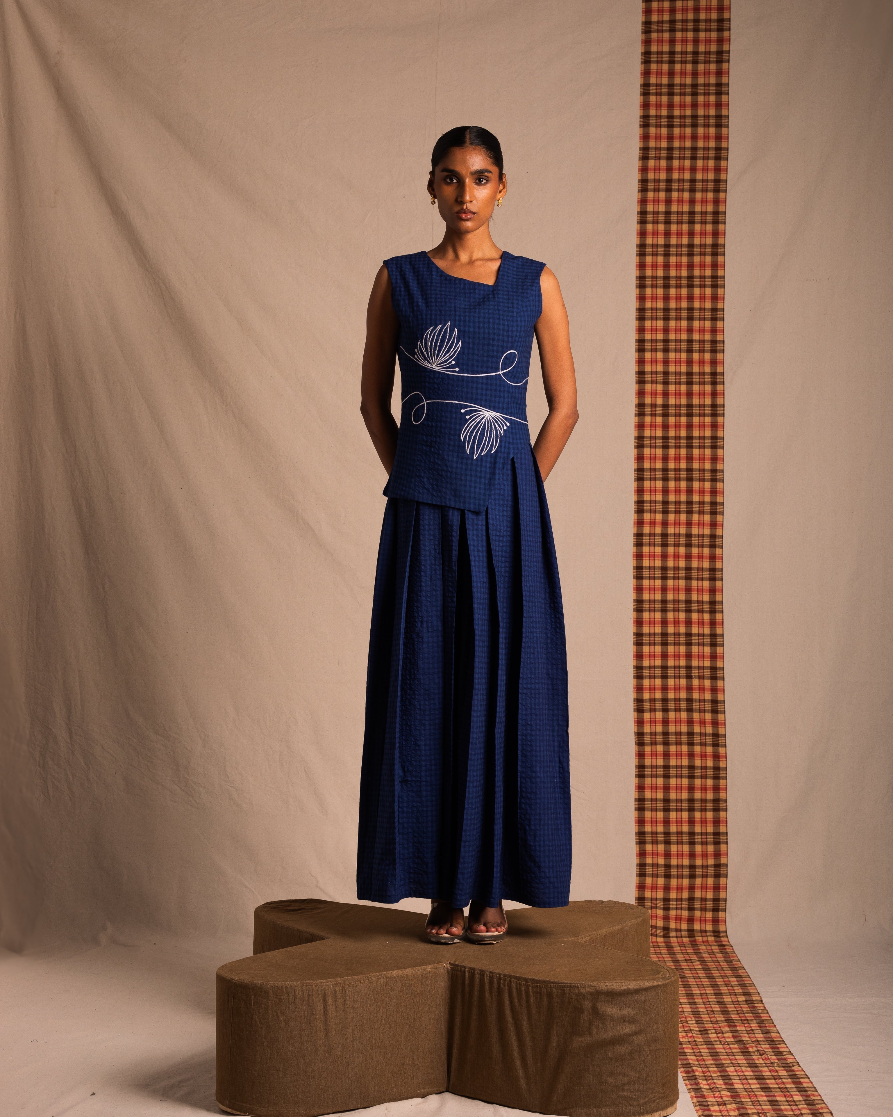 Woman in blue sleeveless top and wide-leg pants with white embroidery, standing indoors.