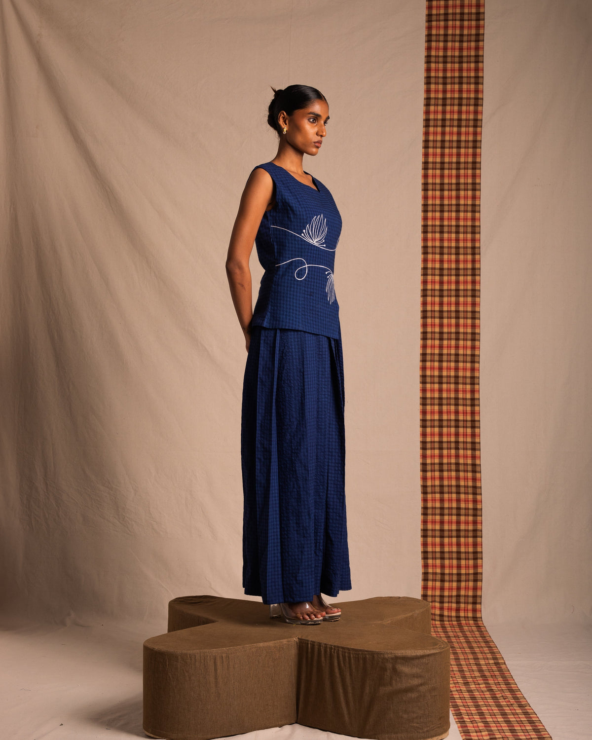Woman in a sleeveless navy blue embroidered top and wide-leg pants, standing on a brown platform in a minimal studio setting