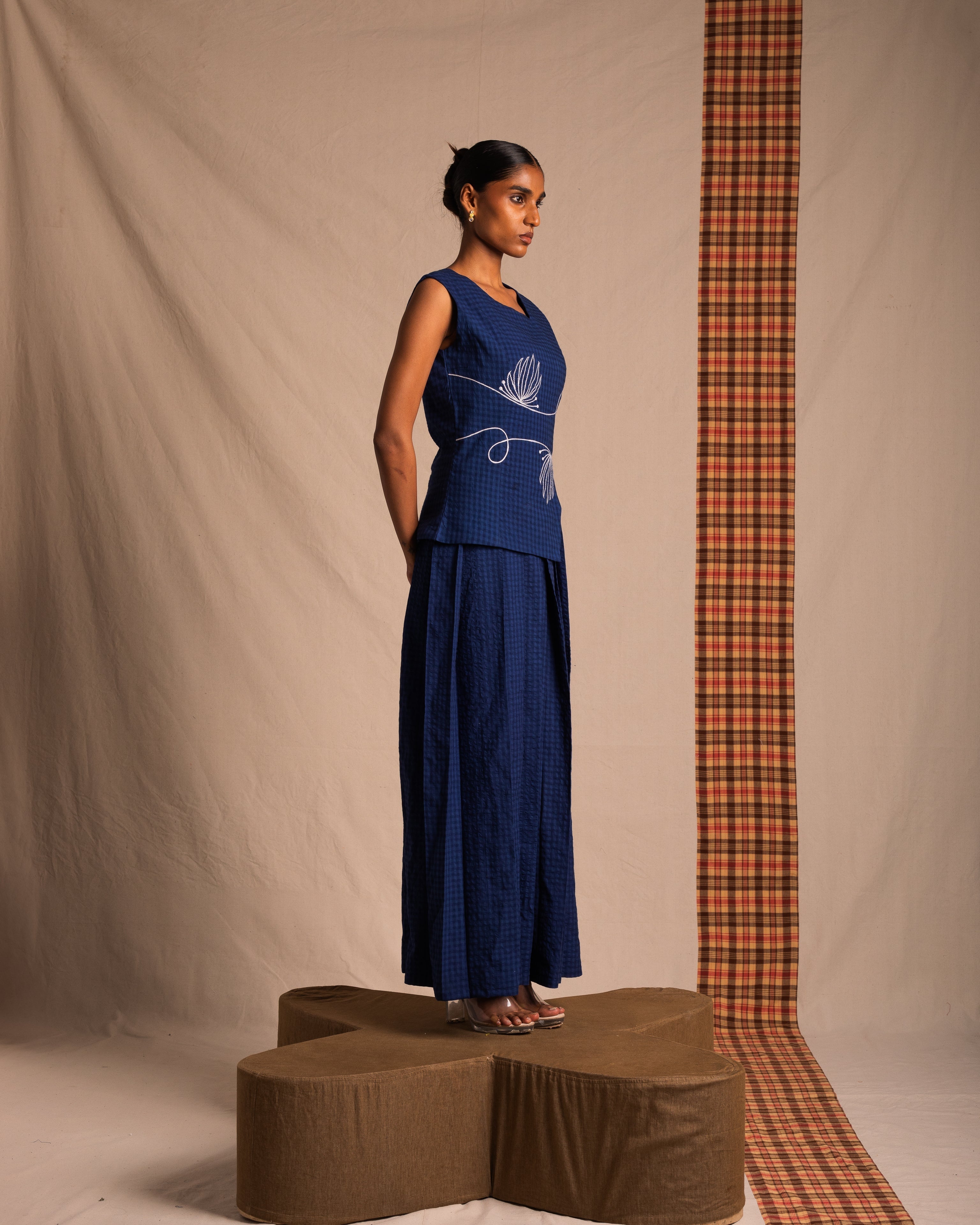 Woman in blue sleeveless embroidered top and wide-leg pants, standing on a brown pedestal