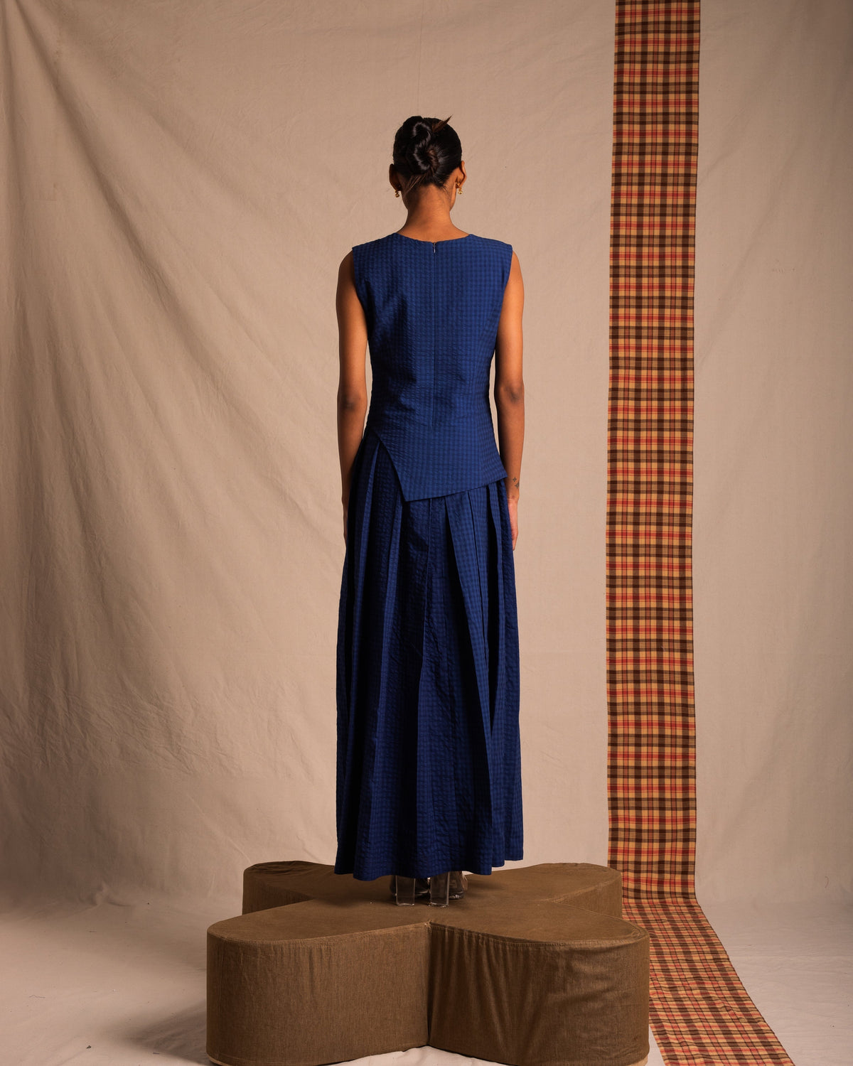 Woman in a sleeveless blue checkered maxi dress, standing on a brown platform with a neutral backdrop